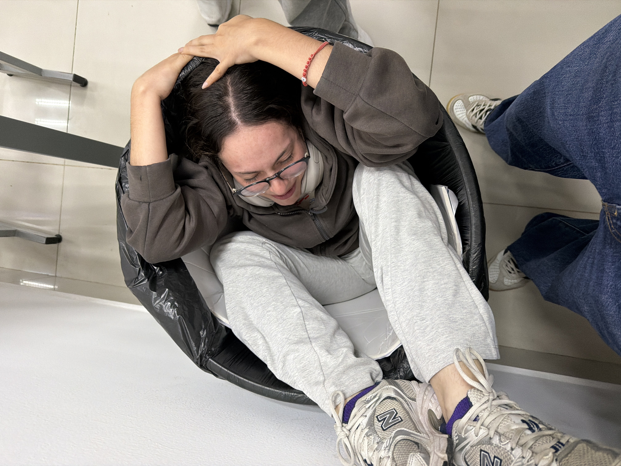 A person is sitting inside a black trash can. The person has brown hair, wears glasses, a brown zip-up hoodie, and grey sweatpants. The person is wearing white and silver New Balance sneakers with purple socks. The person's hands are on their head. Another person wearing blue jeans is standing nearby. The location appears to be indoors with white tiled flooring. Based on the available information, there is no way to confirm this photo was taken in San Andrés Cholula, Mexico.