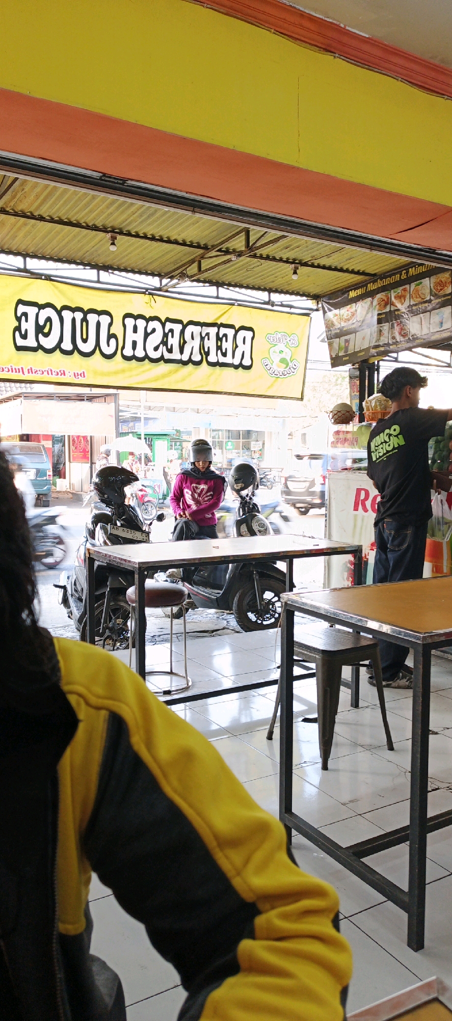 The image depicts the exterior of a juice stand in Sidoarum, Indonesia. A prominent yellow banner with bold, white lettering reads "REFRESH JUICE," partially obscured by reflections and framing. To the right, a menu board displays food and drink options.
In the foreground, a person wearing a yellow and black jacket is visible from the back, their arm resting on a table. Seated on a motorcycle, a person in a pink shirt and headscarf is looking towards the right. Another motorcycle is parked next to theirs.
Behind the motorcycles, a man in a black t-shirt with a green logo that reads "KINGDOM VISION" is interacting with items at the juice stand. He appears to be serving or arranging produce. The environment includes tiled flooring and metal furniture. The overall scene suggests a busy street-side establishment.