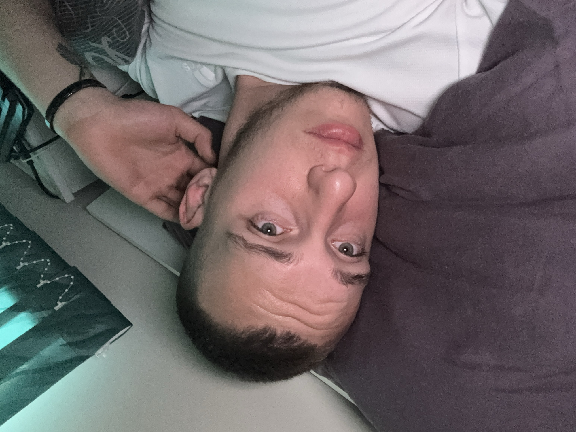 This image, taken in Dobrogled, Bulgaria, features a man lying down, photographed from an overhead perspective, with his head oriented upside down in the frame. The subject appears to be a man in his 20s or 30s, with short dark hair and stubble or a short beard. He is wearing a light-colored, possibly white or light grey, t-shirt, on which a faint Adidas logo can be discerned on the shoulder. His left arm is visible, featuring a dark bracelet or band on the wrist and a tattoo on the upper arm/shoulder area.
He is partially covered by a dark grey or purplish blanket or fabric that stretches across the right side of the frame. The man is looking directly into the camera with an open-eyed, neutral to slightly pensive expression. In the background, to the left, there is a greenish-blue object, possibly a screen or a framed artwork, displaying some white, dotted lines or decorative patterns. The setting appears to be an indoor space, likely a bedroom or living room, with general indoor lighting, suggesting either daytime without direct sunlight or evening with artificial light. The man is actively posing for the photograph.