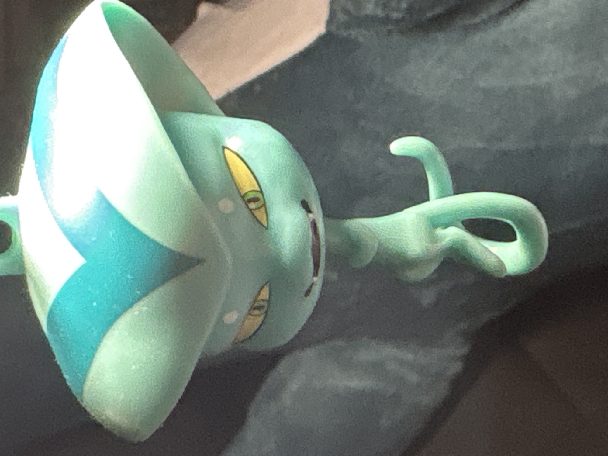A close-up shot of a Suicune Pokemon toy is shown. The toy is mostly mint green and teal. It has two eyes with yellow irises, and a dark purple line for a mouth. The toy's head has a large white crest with teal markings. The toy is sitting on a dark surface with a blurred background. The location is indeterminable from this image.