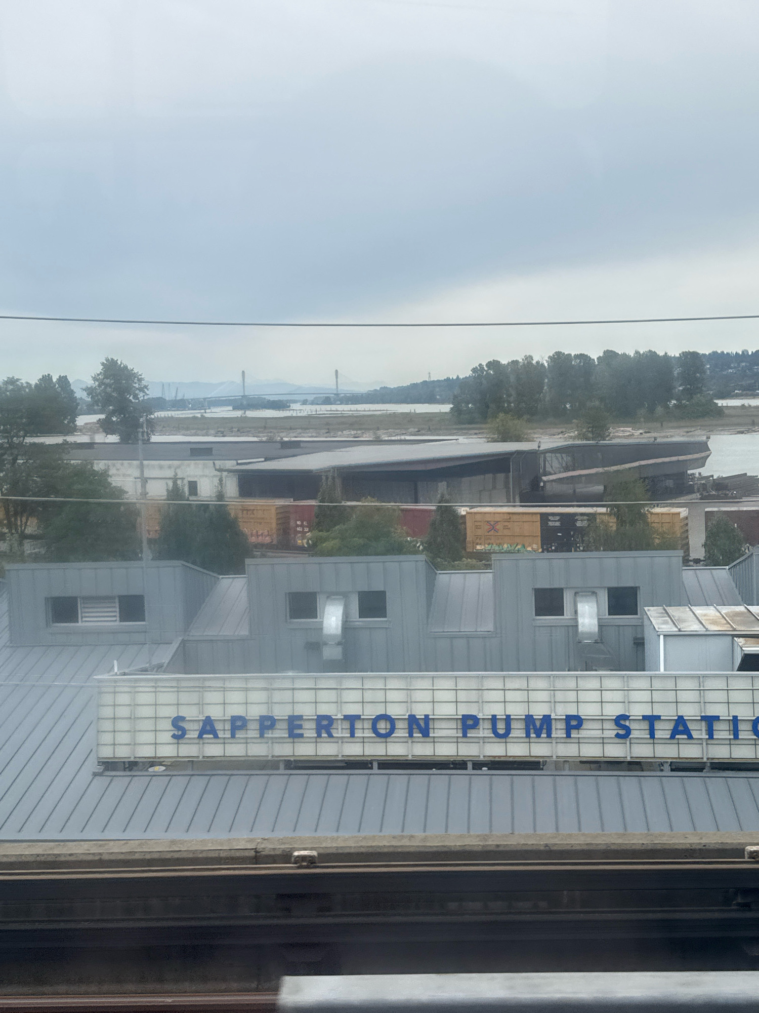 The image captures a view of the Sapperton Pump Station in New Westminster, Canada. The scene is dominated by the industrial architecture of the pump station, characterized by its grey, metal-clad roofs and prominent signage. The words "SAPPERTON PUMP STATION" are displayed in blue lettering against a grid-like white background, mounted above the main structure.
In the background, a body of water, likely the Fraser River, is visible. Beyond the water, a densely wooded shoreline is present, with trees forming a green band against a hazy sky. A suspension bridge with prominent towers can be seen in the distance, spanning the river. The sky is overcast, suggesting a cloudy or foggy day.
In the foreground, a section of a railway track is visible, with a dark, textured surface. The overall impression is one of an industrial waterfront area under a muted sky. No people are visible in the frame.