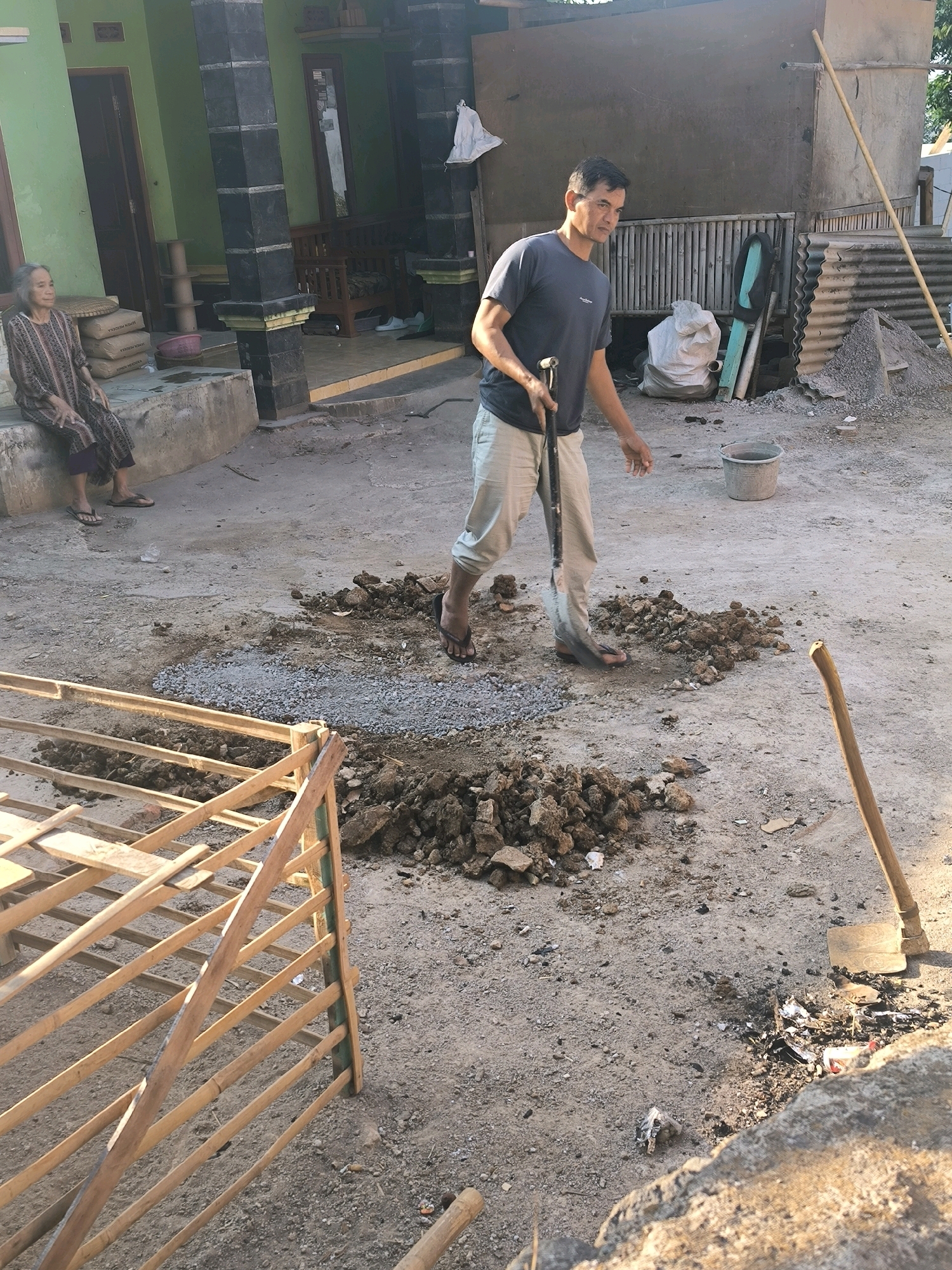 In the city of Cilengkrang, Indonesia, a man is seen holding a shovel and a cane in an outdoor courtyard, with a woman sitting on a raised concrete platform to his left. The man, wearing a dark t-shirt and light-colored pants, appears to be working with the soil and gravel scattered on the ground. A wooden structure, possibly part of a construction project, is visible in the foreground. The environment suggests a construction or renovation site, with various building materials and tools present, including bags of cement, corrugated metal sheets, and buckets. The scene is illuminated by natural daylight, casting shadows across the ground.