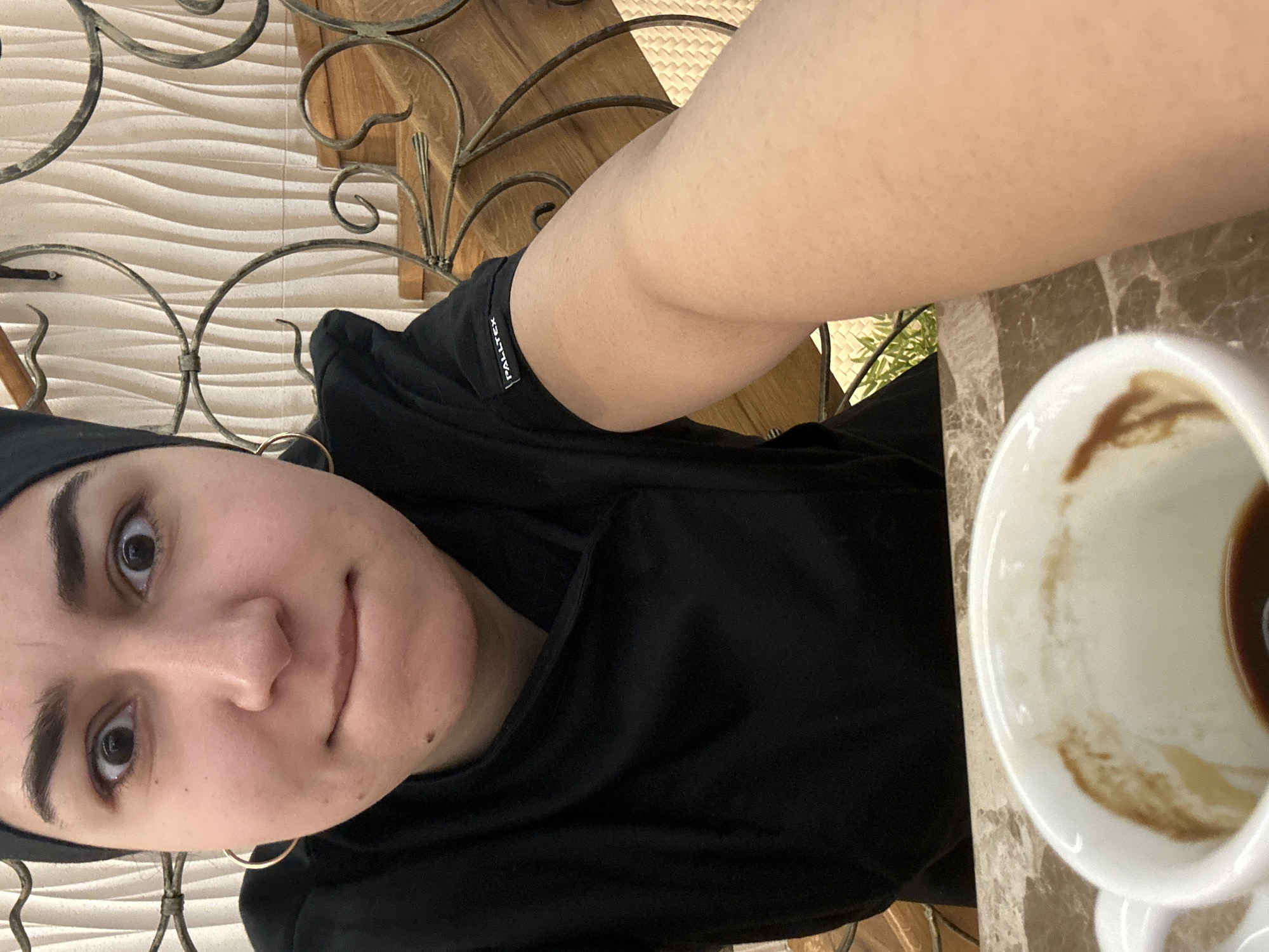 A selfie of a person wearing a black headscarf, a black sweatshirt with the label "PALLTEX" visible on the sleeve, and gold hoop earrings. They are sitting at a table with a half-empty white coffee cup on it. The table has a marble-like pattern. Behind them is a decorative iron structure and a wall with a wavy texture.
The location is likely Burgas, Bulgaria.