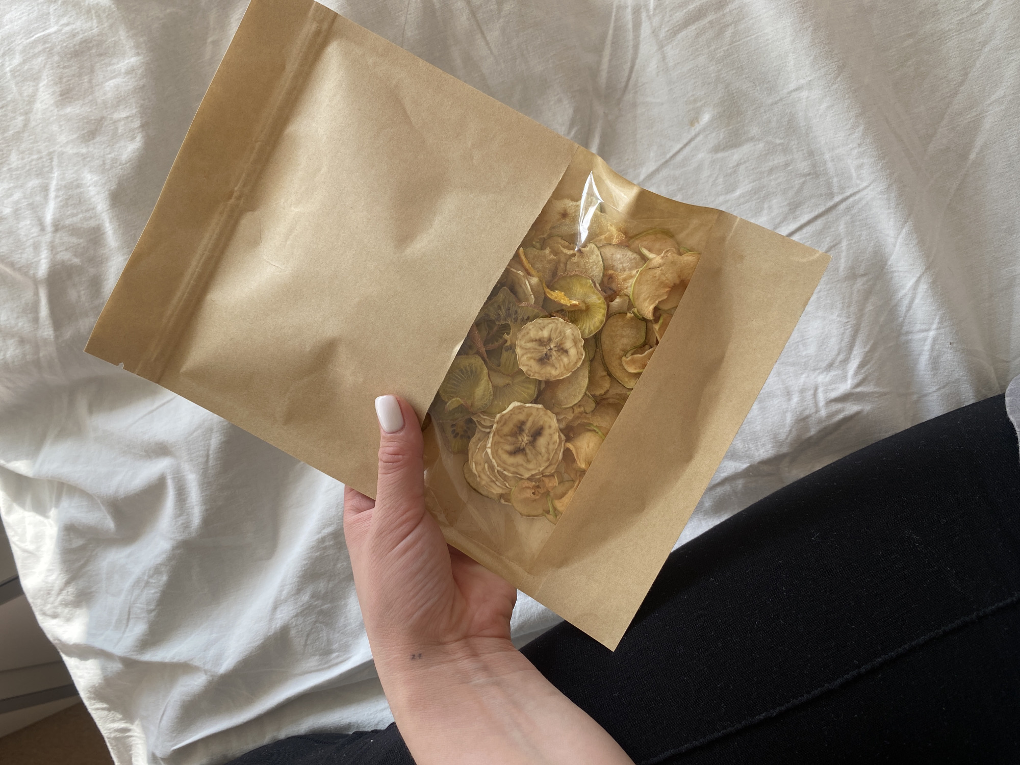 A person holds a partially open brown paper bag of dried fruit snacks. The bag contains dried slices of banana and kiwi. The person has fair skin, a white fingernail, and a tattoo on their wrist. They wear black clothing. The background is a wrinkled white sheet. The image suggests an indoor setting. The city of Perm, Russia, is not discernible from the image.