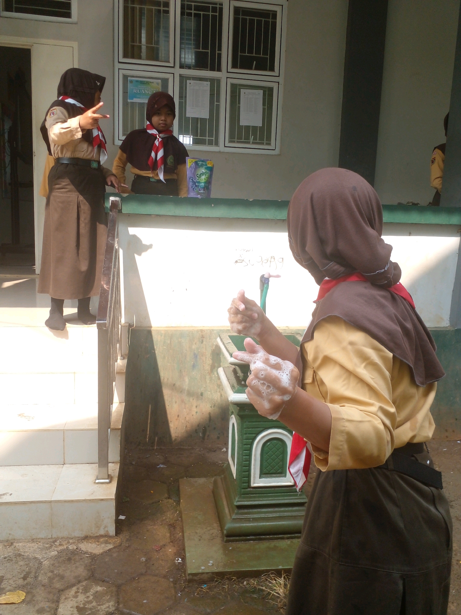 The image shows two young girls in uniform at what appears to be a school in Banyuputih, Indonesia. The girl on the right is washing her hands with soap and water from a faucet, her hands covered in foam. She is wearing a brown headscarf, a tan shirt, and a dark brown skirt. The girl on the left is also in uniform, wearing a brown headscarf, a tan shirt, and a dark brown skirt. She is standing on a set of steps, looking towards the other girl and making a "peace" sign with her fingers. A package of Sunlight dish soap is visible on a counter behind them. The environment appears to be outdoors, with a tiled walkway and a dirty, paved area with some puddles. The building behind the girls has windows with barred panes and posters or notices displayed inside. The overall scene suggests a moment of hygiene practice in an educational setting.