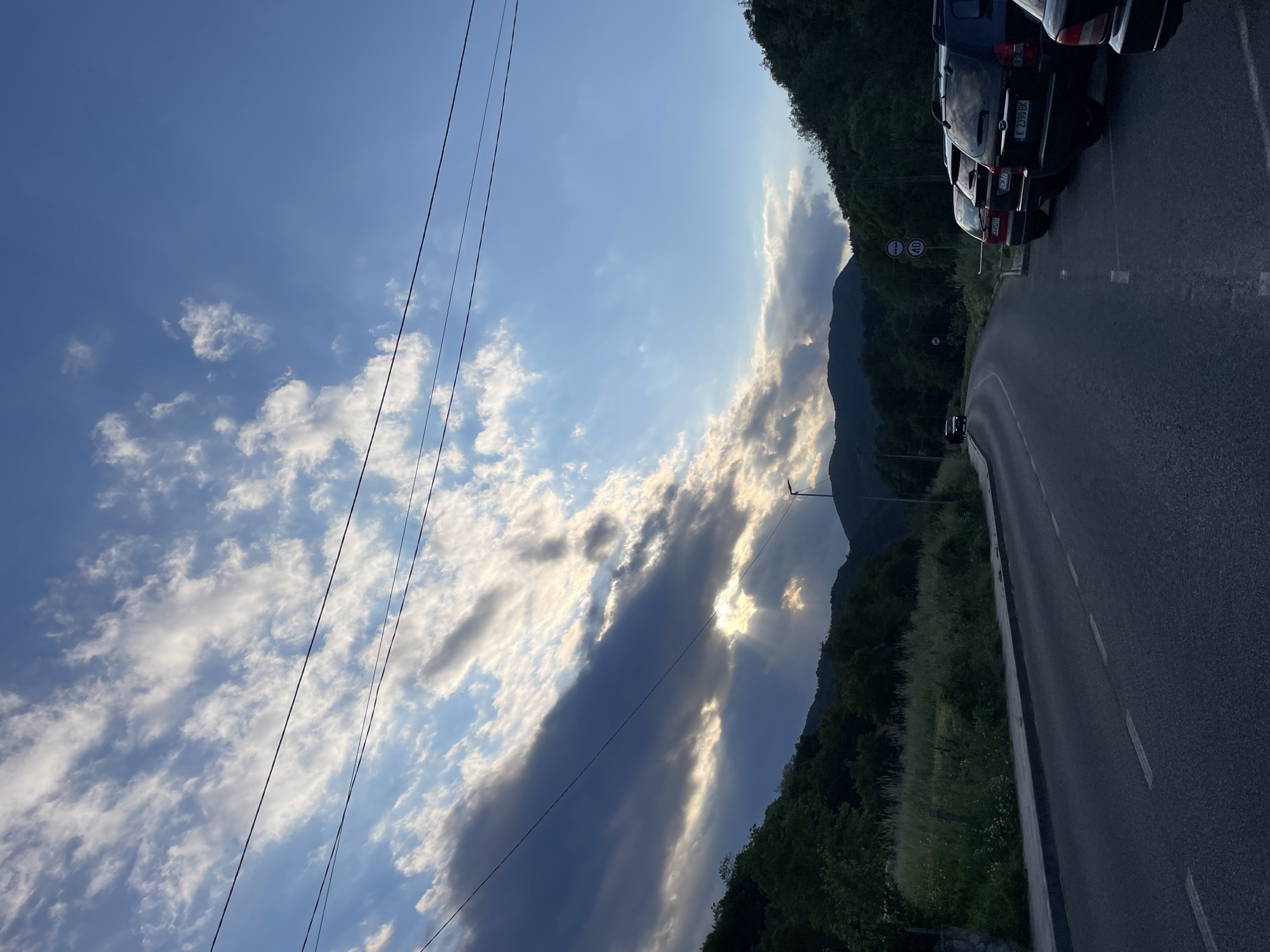 The image shows a road winding along a mountainside in Zlatograd, Bulgaria. The sky is filled with scattered clouds, some obscuring the sun. Power lines run along the left side of the frame. A row of dark-colored cars is parked on the shoulder of the road. A speed limit sign is visible near the cars. Vegetation covers the mountainside.