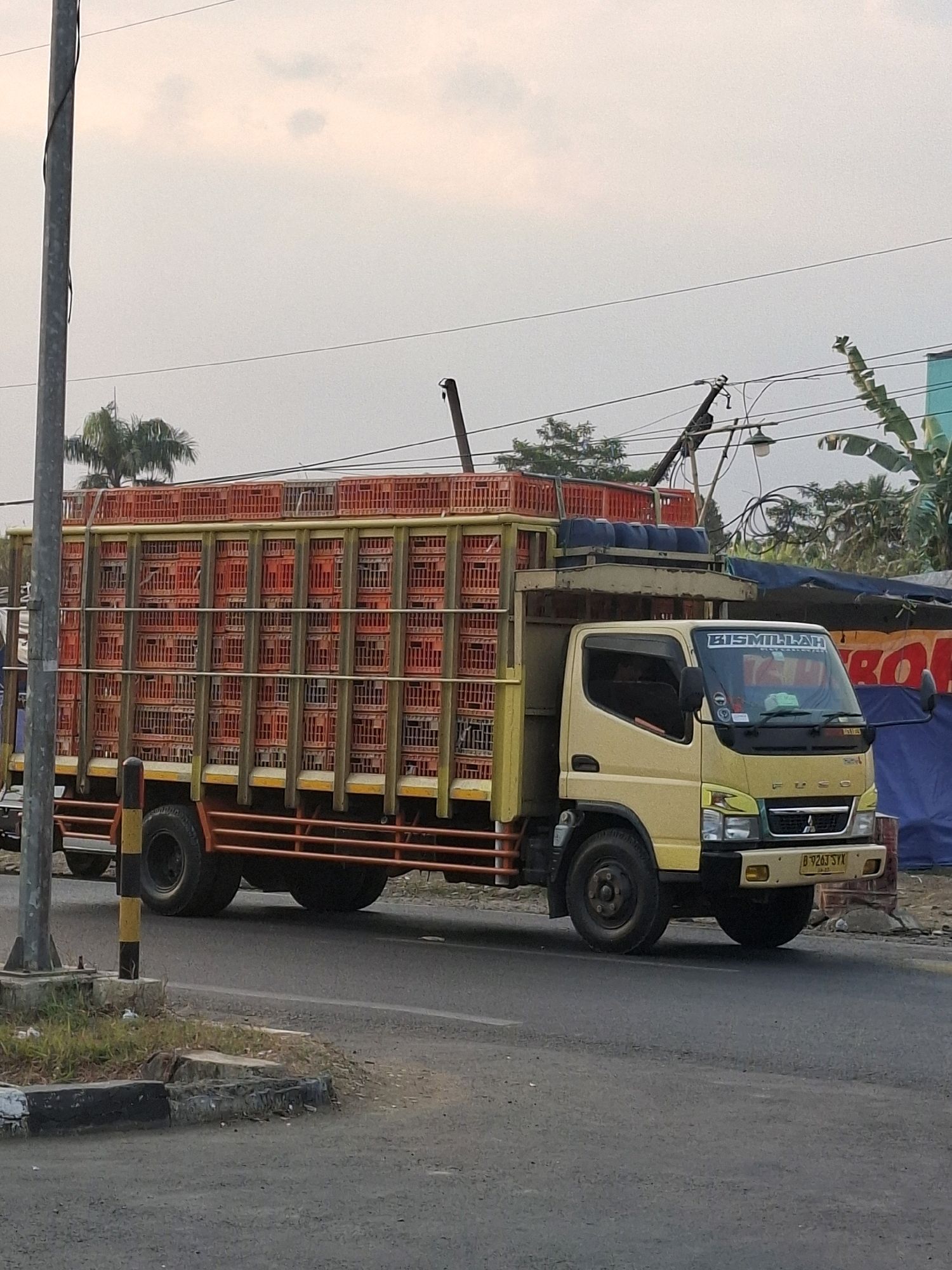 A yellow Mitsubishi Fuso truck, its cargo area filled with stacked orange plastic crates, drives on a paved road in what appears to be a semi-urban or rural area. The truck has a "Bismillah" decal on its windshield. The sky is overcast, suggesting either early morning or late afternoon light. A utility pole stands to the left of the truck, and vegetation, including palm trees, is visible in the background. There are other structures and trees in the distance, and a blue tarp is present on the right side of the image. The license plate on the truck is partially obscured but appears to read "B 9263 SYX". The truck is traveling from left to right.