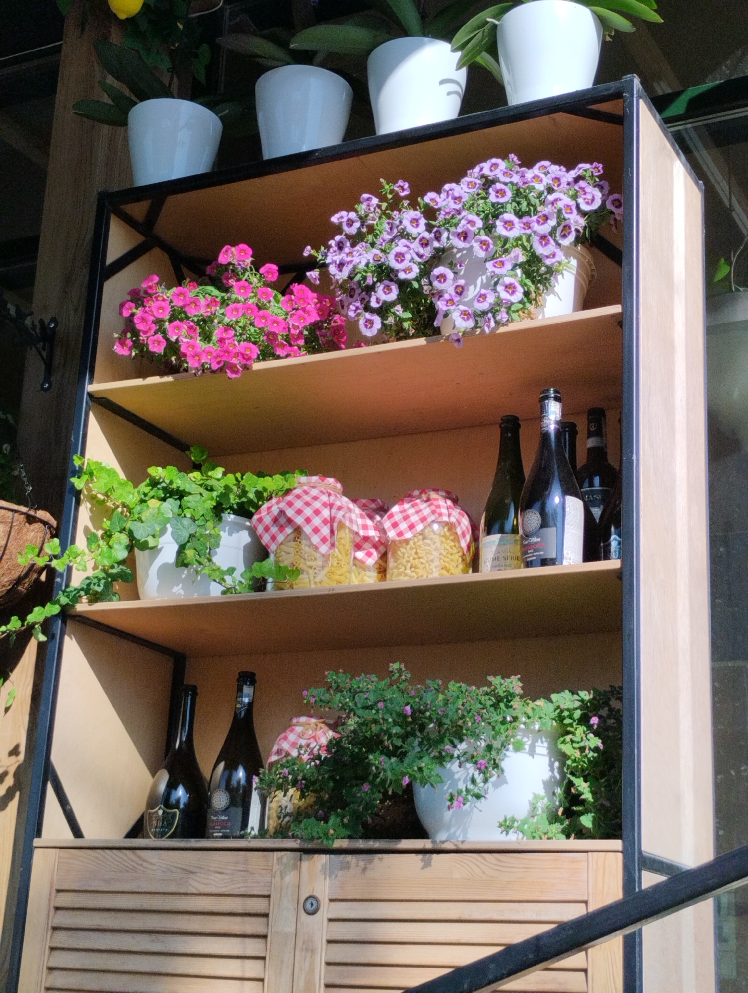 The image shows a tall, multi-tiered wooden and metal shelving unit filled with plants and bottles. The top shelf displays two white pots containing small pink and purple flowers. Below that, a shelf holds a potted green ivy plant and two glass jars filled with pasta, covered with red and white checkered fabric. The next shelf down contains several dark glass bottles, possibly wine or spirits. The bottom shelf has two dark glass bottles and a potted plant with small purple flowers. The shelving unit is placed indoors, with natural light illuminating the scene. The lower portion of the unit has wooden louvered cabinet doors. The overall impression is of a decorative display, possibly in a home or a cafe. The location of Kostanay, Kazakhstan is not visible in the image.