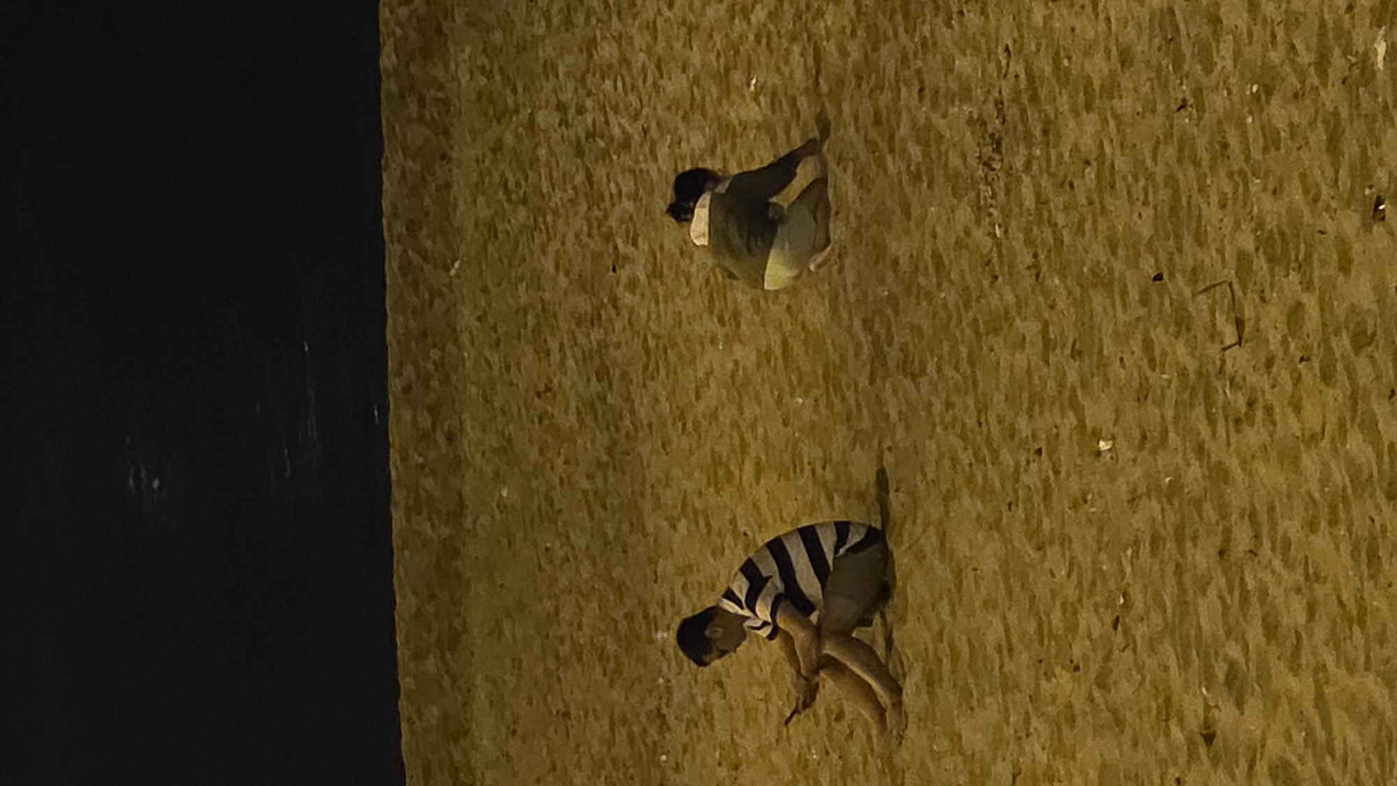 A nighttime, low-angle shot depicts two individuals scaling a textured yellow wall. The upper figure is partially obscured by the wall's edge, wearing a dark top and light-colored bottoms, and appears to be looking down. Below, a second individual, wearing a black and white striped shirt and shorts, is actively climbing. The wall appears to be part of a structure situated next to a dark body of water. This image was taken in Nha Trang, Vietnam.