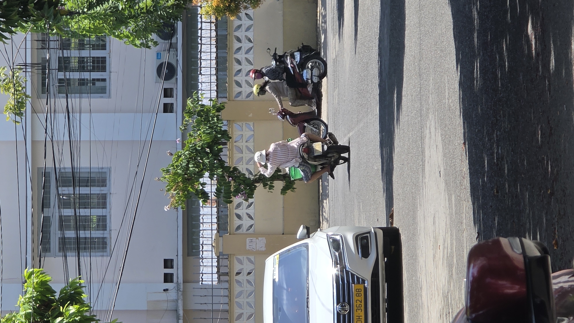 The image depicts a street scene in Nha Trang Ward, Vietnam. Three people are riding motorbikes along the street. A white vehicle is parked along the street. The license plate appears to read "DH-362.88". The background includes buildings, trees, and telephone wires.