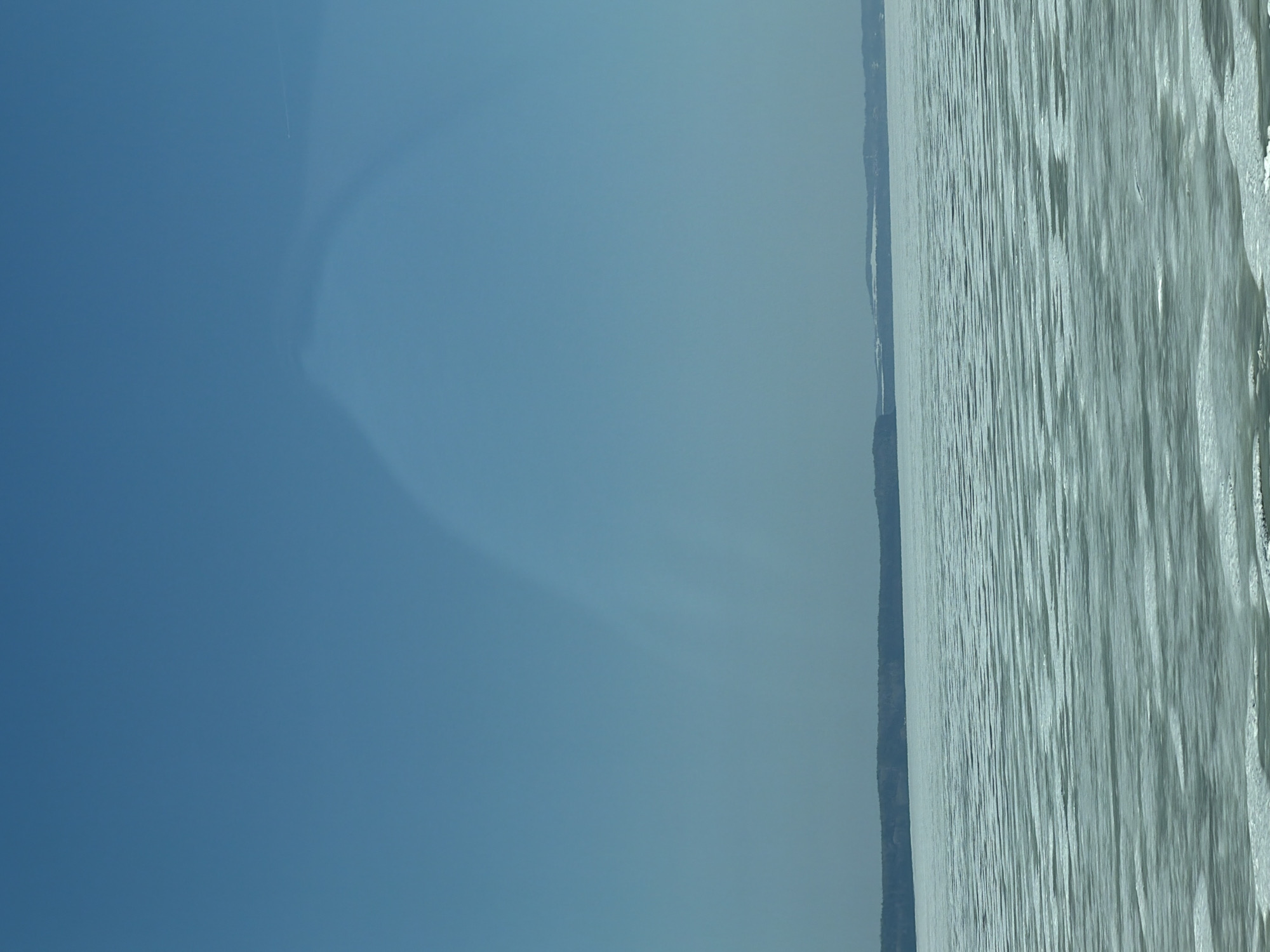 The image depicts a body of water with a visible shoreline in the distance. The sky is clear and blue. The photo was taken near the city of Chaikovsky, Russia.