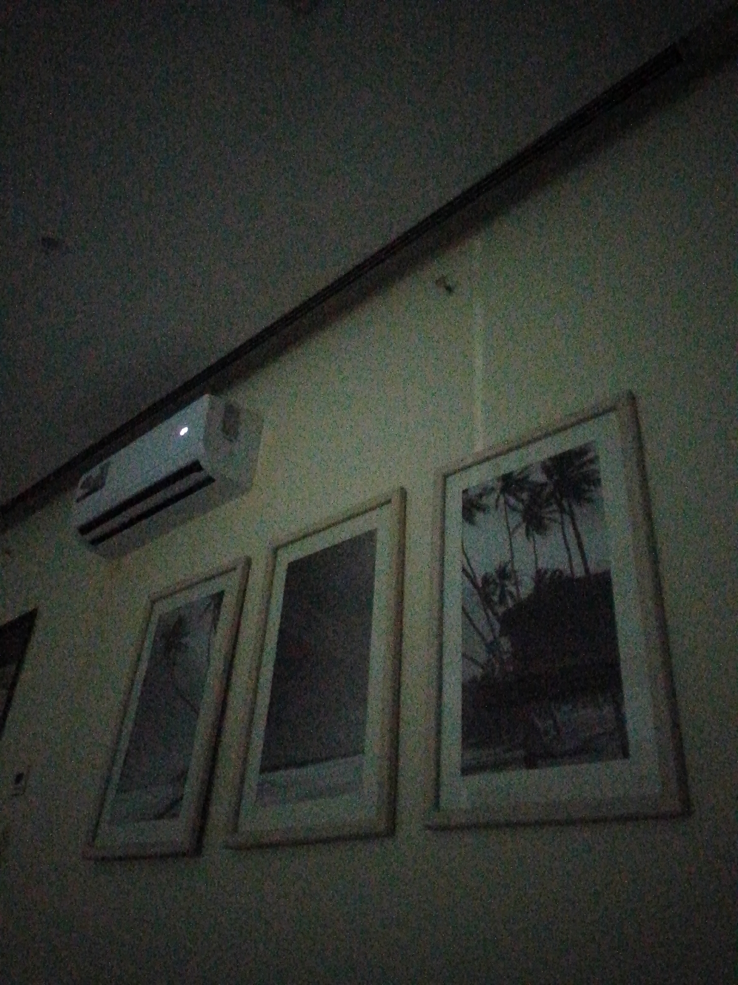 Indoor scene. A white air conditioning unit is mounted high on a wall above three framed pictures. The picture on the right depicts palm trees. The image is noisy and dark, and the location, Pedro González, Venezuela, cannot be determined from the image.