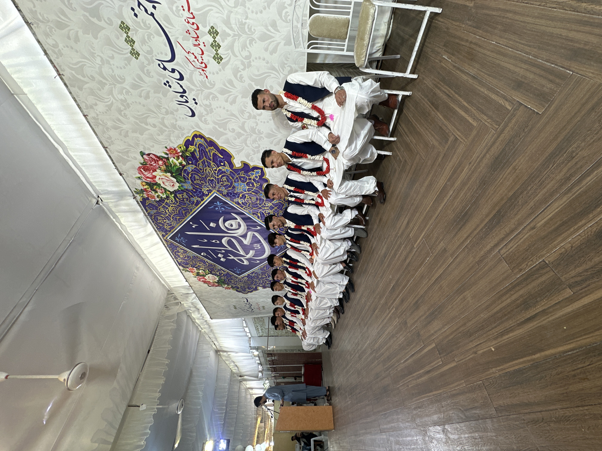The image depicts a group of at least ten young men seated in a row on white chairs in what appears to be an indoor event space, possibly a hall or large tent. They are uniformly dressed in white salwar kameez with dark waistcoats and garlands of flowers. A man stands behind a podium towards the end of the row, facing the seated men. The hall is decorated with white fabric and patterned banners with floral designs and text in Urdu or Arabic. The floor is made of brown wooden panels. The location is in Quetta City Tehsil, Pakistan.
