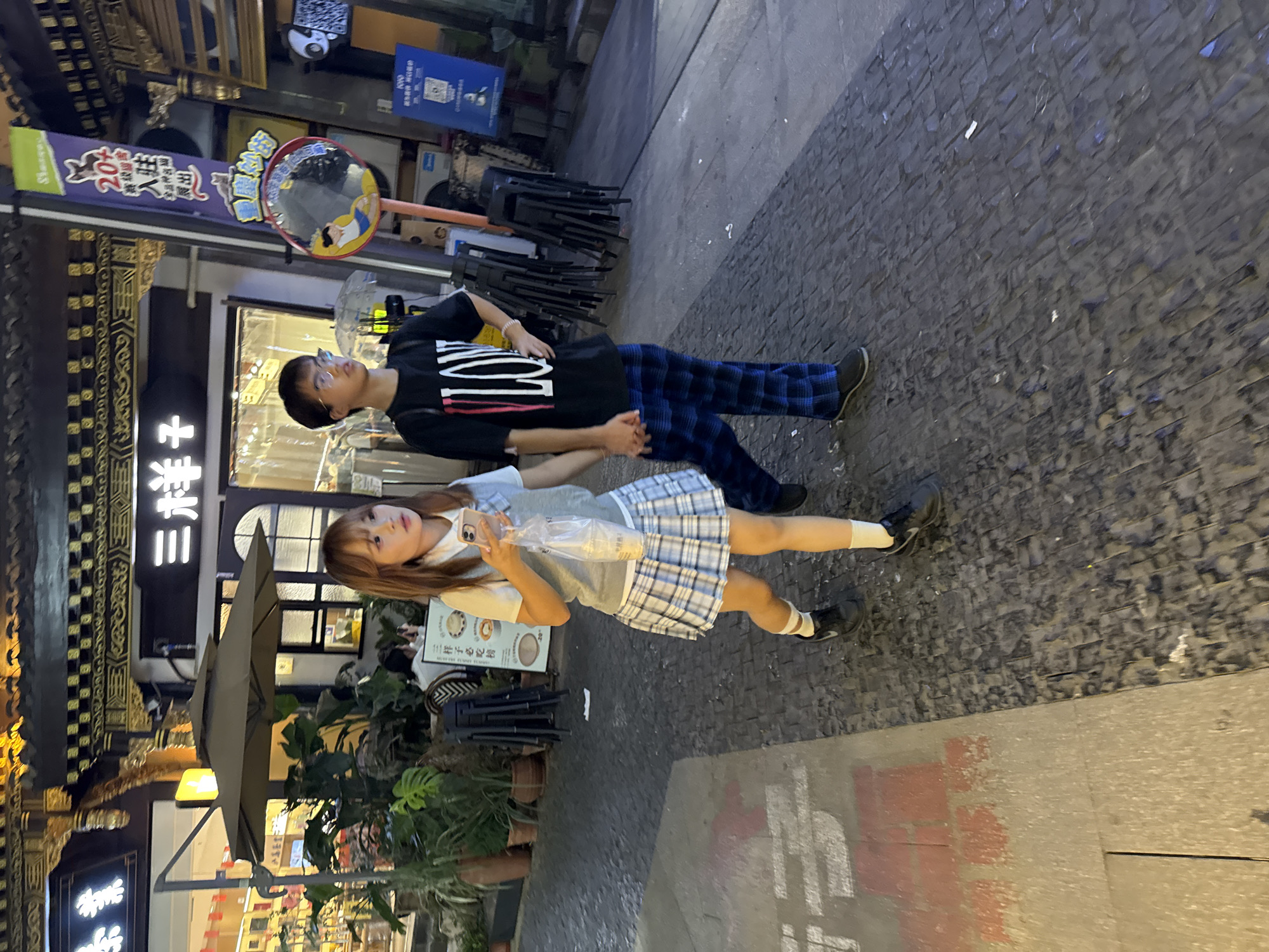 In Yuzhong District, China, a young couple walks hand-in-hand down a cobblestone street at night. The female, on the left, wears a gray vest over a white shirt and a blue and white plaid skirt, with white socks and black sneakers. She holds a plastic bag containing a drink and her phone. The male, on the right, is dressed in a black t-shirt with a white and red "VLONE" graphic and blue and black plaid pants, also wearing dark shoes. They are walking away from the camera. The background features traditional Chinese architecture with ornate gold trim and neon signs, including a banner with Chinese characters and a cat logo. An outdoor seating area with plants and umbrellas is visible to the left, and stacks of chairs are present to the right. The street is paved with dark cobblestones, with a lighter-colored concrete path on the left side of the image.
