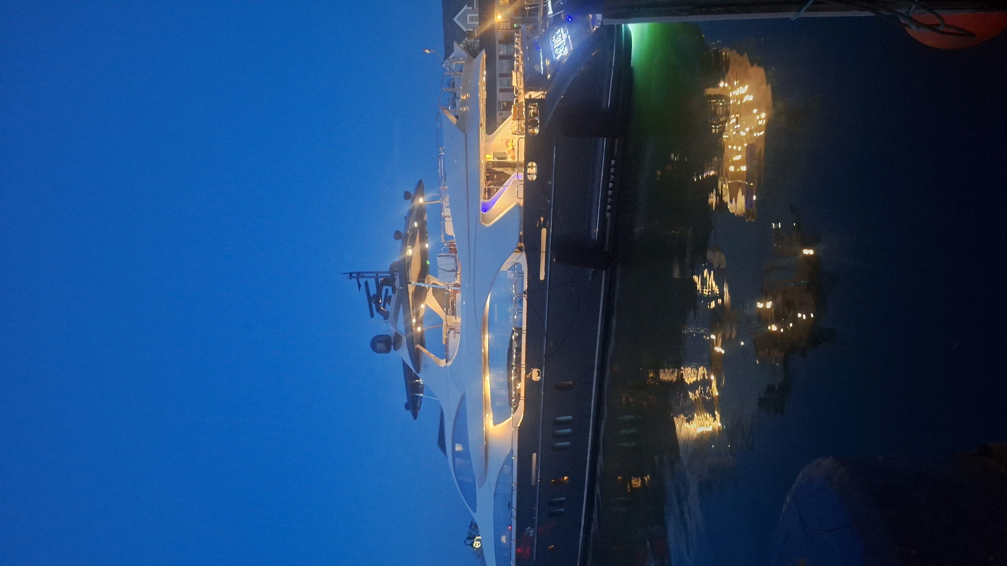 The image shows a large, dark-hulled yacht docked at night in Nantucket, United States of America. The yacht is illuminated by interior lights, casting a warm glow and reflecting on the dark water. The sky is a deep blue, indicating twilight or night. The reflection of the yacht and its lights is visible on the water's surface. In the background, there are dimly lit buildings with lights on, suggesting a harbor or waterfront area. There are no people visible in the image. The yacht has a sleek, modern design with multiple levels and windows. The name "NURORA" is visible in illuminated letters on the stern of the yacht.