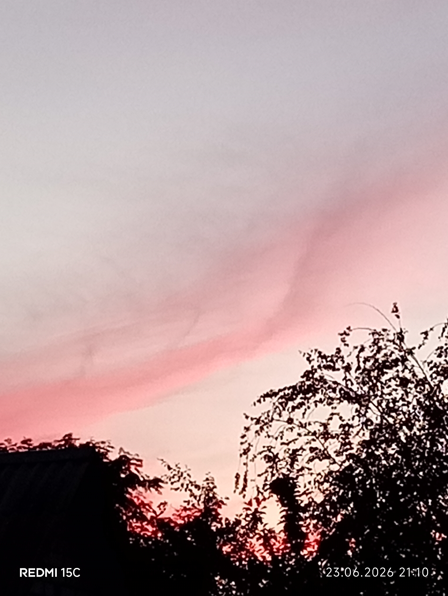The image depicts a sunset sky with hues of pink and grey. Silhouetted against the sky are the tops of trees and the roof of a building. The bottom left corner displays the text "REDMI 15C," and the bottom right corner shows the date and time "23.06.2026 21:10." The scene is devoid of any people or discernible human activity, and the location is identified as Mykolaivka, Ukraine.