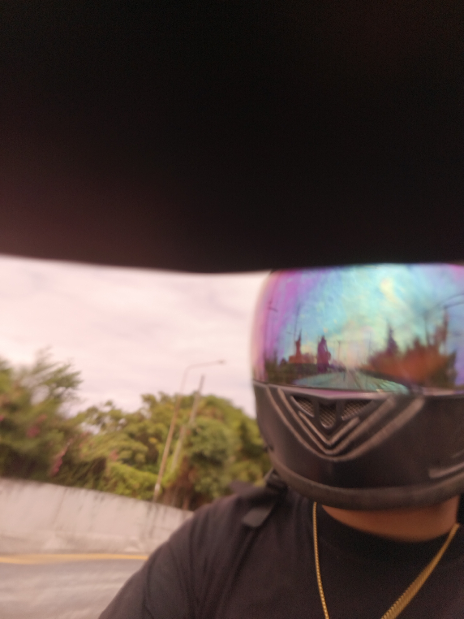 The image shows a first-person perspective of someone riding a motorcycle in Denpasar, Indonesia. The rider is wearing a black t-shirt and a helmet with a reflective visor that displays a colorful, iridescent sheen. The visor reflects the surrounding environment, including trees, a street, and buildings that appear to be temples or ornate structures. The sky is overcast with a light pinkish hue. The road is visible in the foreground, with a yellow line marking the lane. A concrete barrier is also present alongside the road, with lush green trees and foliage in the background. The overall scene suggests a ride through a suburban or semi-urban area of Denpasar, possibly near a cultural or religious site given the reflected temple-like structures.
