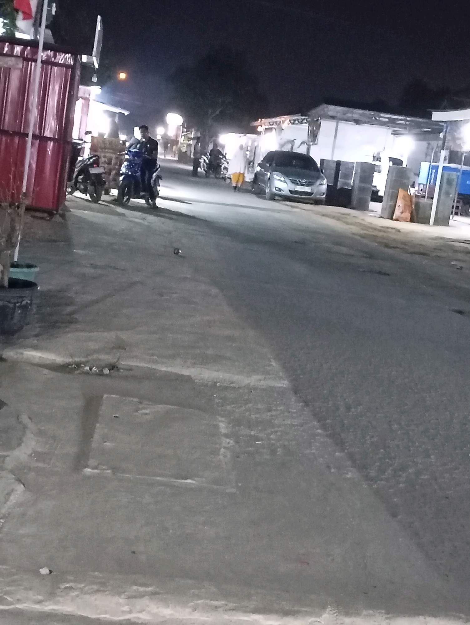 The image captures a nighttime scene on a street in Leman Benawi, Indonesia, likely in a semi-urban or commercial area. The foreground features a paved road and a sidewalk or elevated walkway with some debris. To the left, a red metal structure is partially visible, possibly a building facade or stall.
In the mid-ground, several motorcycles are parked and one is being ridden by a person wearing a dark shirt. Further down the street, a silver sedan is parked, and a person in a light-colored garment, possibly a hijab, is walking away from it. Structures that appear to be shops or stalls with corrugated roofs and lighting are present in the background, illuminated by artificial light. The overall atmosphere is one of activity on a street at night, with vehicles and pedestrians present. The lighting conditions suggest it is dark outside, with streetlights and lights from the establishments providing illumination.