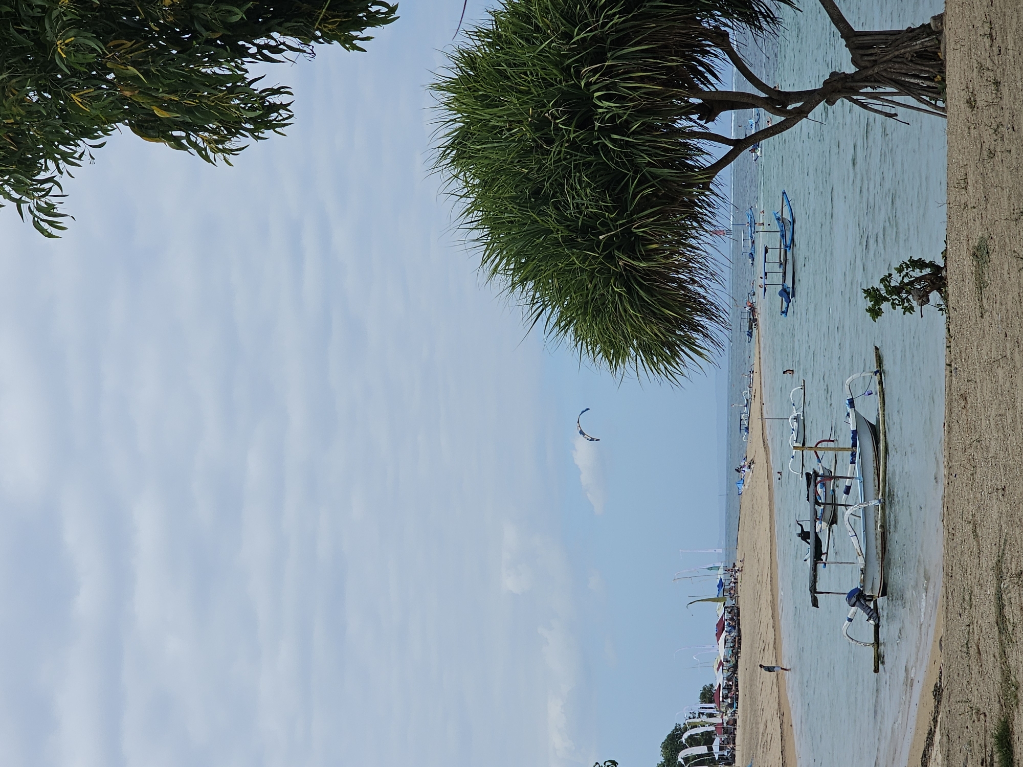 A serene beach scene unfolds with a sandy shore and calm, turquoise waters. Several traditional Balinese fishing boats, known as jukung, are moored near the beach, some with outboard motors visible. In the distance, a kite is visible in the sky, suggesting a breezy day. The shoreline is lined with beach umbrellas and lounge chairs, indicating a popular spot for relaxation and recreation. Palm trees and other lush vegetation frame the view, adding to the tropical ambiance. The sky is overcast with soft, diffused light, creating a peaceful atmosphere. The image captures a typical day at a beach in Denpasar, Indonesia, a popular tourist destination known for its beautiful coastlines and vibrant culture.