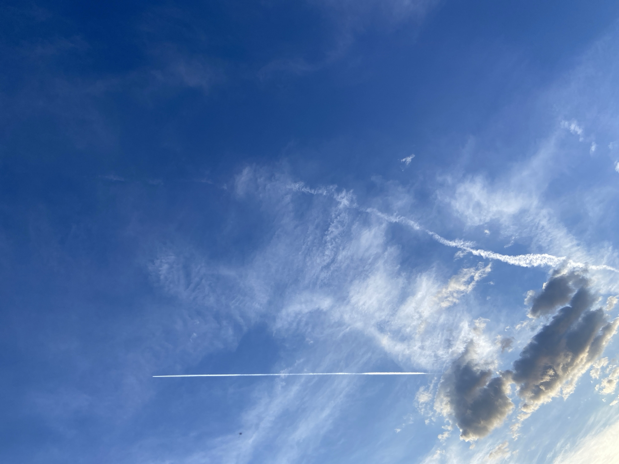 The image shows a clear blue sky with various types of clouds. A distinct contrail stretches horizontally across the frame, left by an aircraft. Another thinner, partially dissipated contrail is visible higher up. Some cumulus clouds are present in the lower right portion of the image. The location is Unknown, Unknown.
