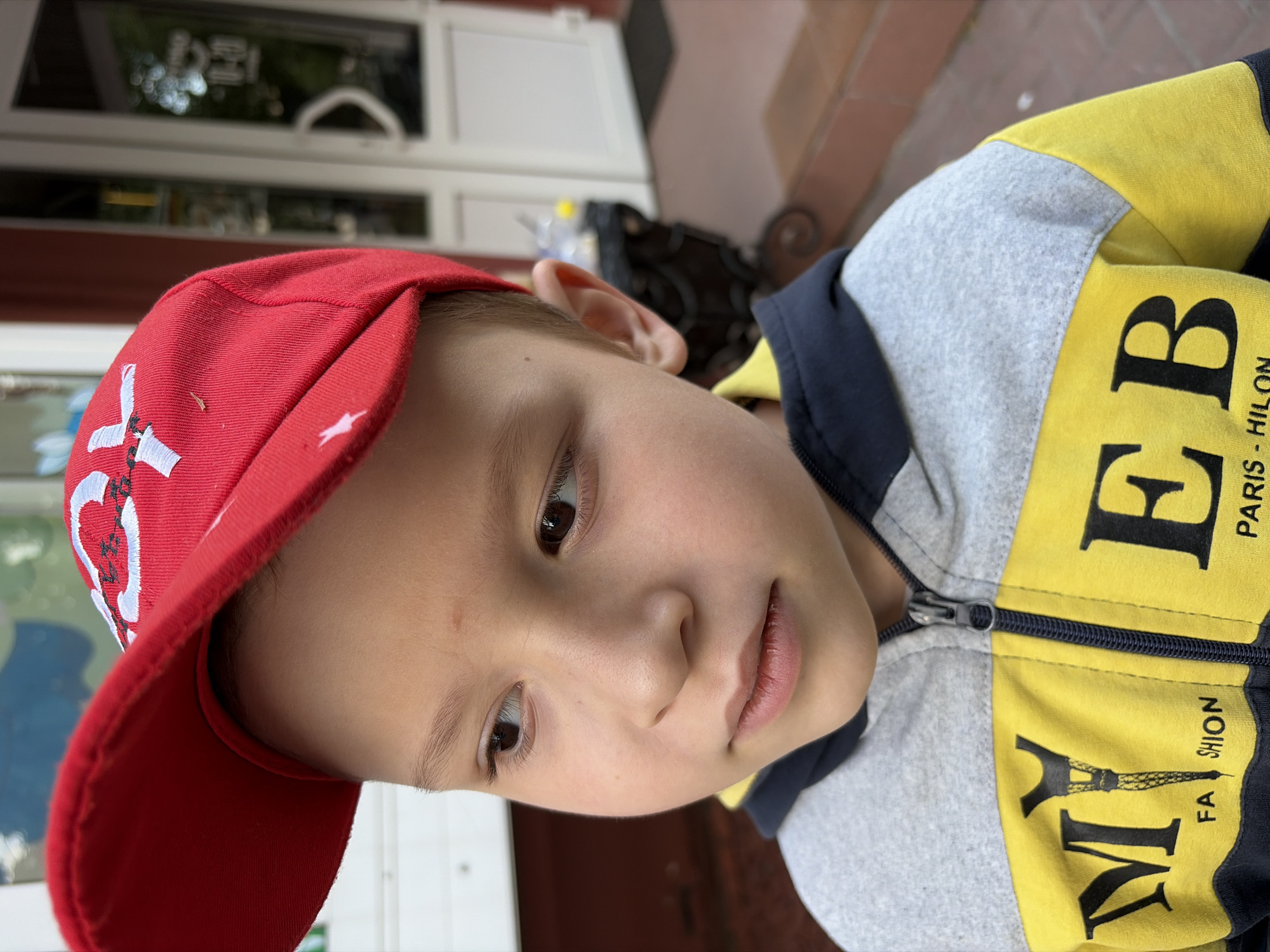 The image shows a close-up of a young boy wearing a red baseball cap and a grey and yellow zip-up hoodie. The boy is looking to his left, with his eyes and part of his face in focus. The background is blurred but appears to be an outdoor setting with a building and some greenery. The boy's hoodie has the words "MY" and "EB" printed in large black letters on the yellow portion, with smaller text below that reads "FA SHION" and "PARIS - HILON". The image is taken from a slightly low angle, looking up at the boy. There is no explicit information in the image to pinpoint the location to Chernihiv, Ukraine.