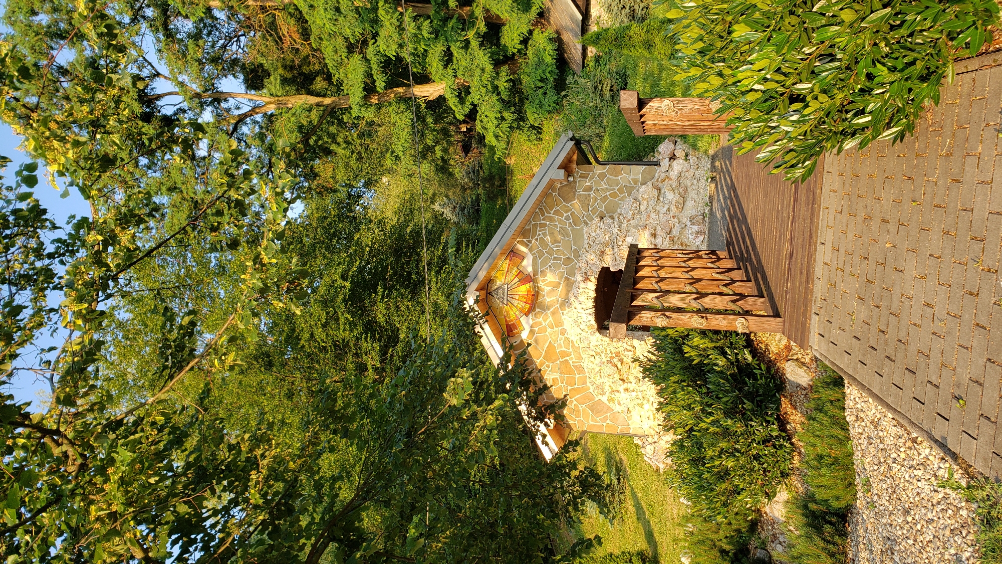 The image depicts a small stone building with a wooden bridge leading to its entrance. The building has a light-colored stone facade and a pitched roof. A colorful mosaic, resembling a sundial, is installed on the gable end of the building. The bridge is made of wooden planks and has wooden railings. The building is surrounded by lush green vegetation, including bushes and trees. The ground in front of the bridge is paved with interlocking concrete blocks. The scene is bathed in the warm light of what appears to be late afternoon or early morning sun. This structure is located at the Kalvária (Calvary) in Prešov, Slovakia.