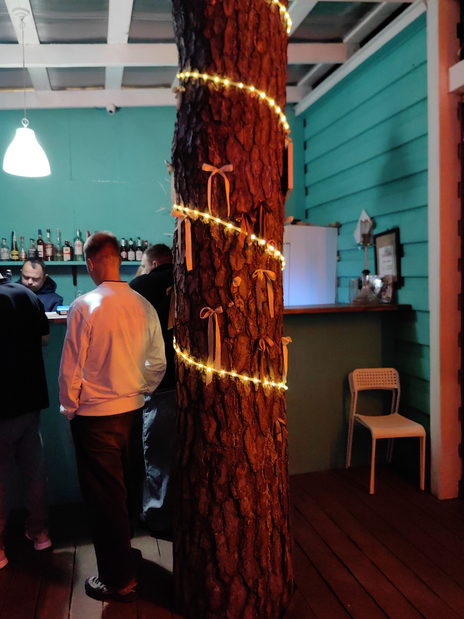 The image depicts an outdoor, possibly covered, seating area. A large tree trunk wrapped with a string of yellow lights and orange ribbons is prominently featured in the foreground. Behind the tree, a bar area is visible with several bottles on shelves and a bartender. Three individuals are present; one is standing at the bar, another is facing away from the camera with their back to the bar, and a third person's back is visible as they stand further back. A white chair is positioned near the bar. The walls are painted teal and white, and the floor is wooden. A white pendant light hangs from the ceiling. The scene appears to be a casual gathering at a bar or restaurant.