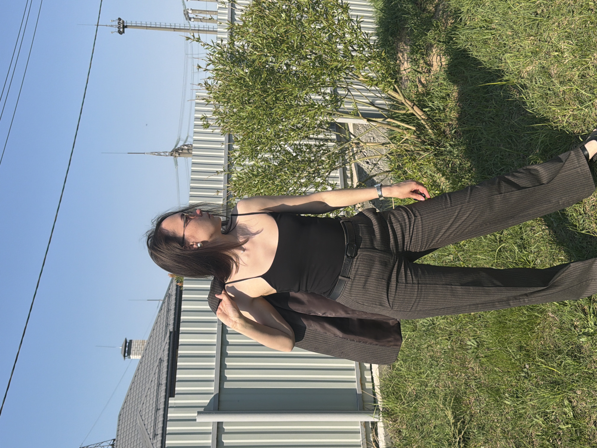 A woman wearing a black tank top and brown pinstripe pants stands on a grassy area. She wears a watch on her left wrist and carries a brown jacket over her left shoulder. Behind her is a building with a gray roof and light gray siding. In the background, power lines and towers are visible against a clear blue sky. The location appears to be Chaikovsky, Russia, based on the architecture and environment.