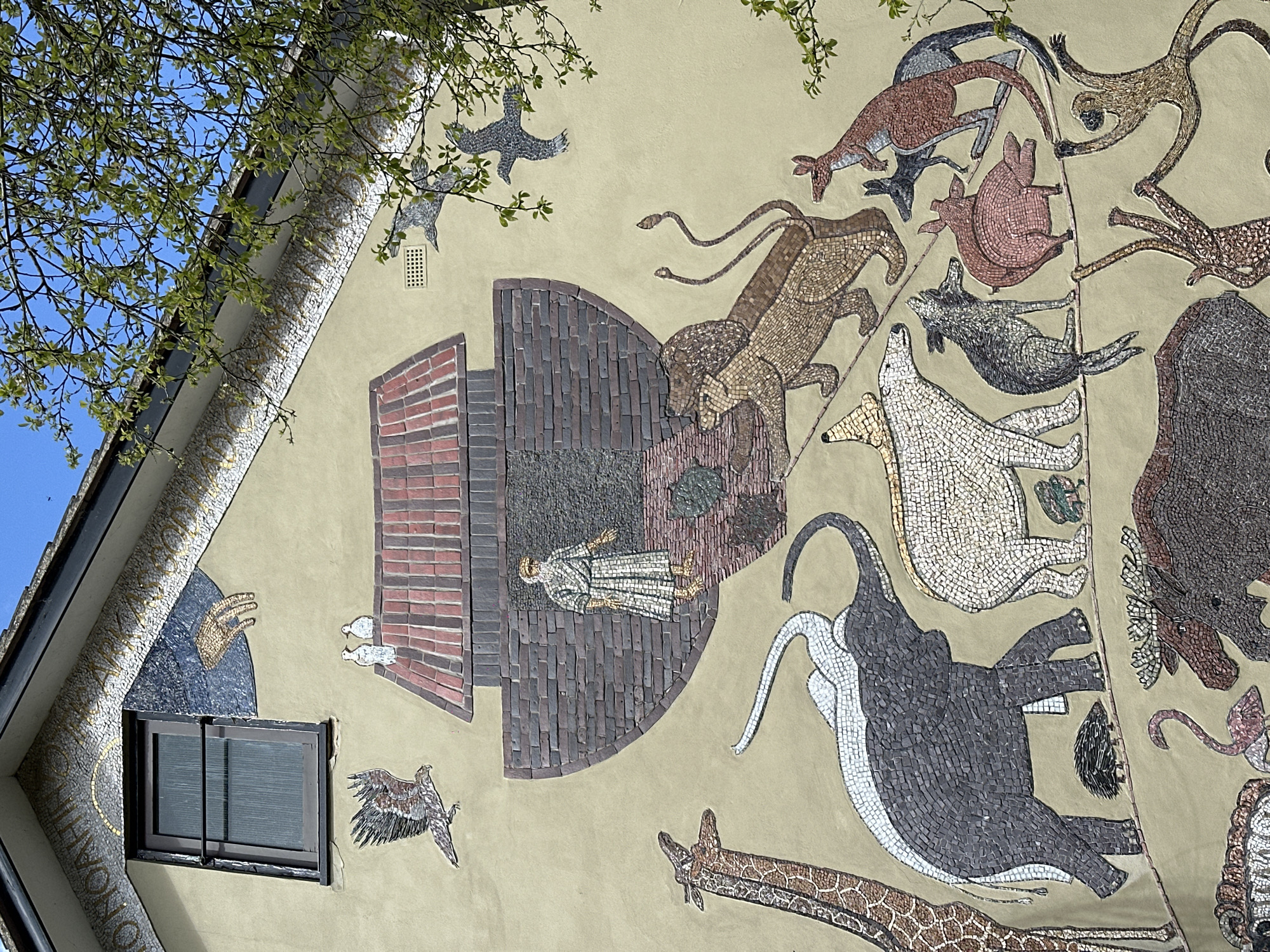 This image depicts a large, detailed mosaic mural on the light beige stucco facade of a building in Maldon, United Kingdom. The mural illustrates the story of Noah's Ark.
In the upper left quadrant, a dark-framed window is visible. Above it, a gold mosaic inscription, partially visible as "NOAH," is set against a white mosaic background. To the right of the window, a dark blue semi-circular mosaic features a golden mosaic hand descending from above. Below the window, a mosaic of a hawk or eagle in flight points left.
Centered on the facade, a large mosaic representation of Noah's Ark features a reddish-brown tiled roof and darker brown brick-like hull. An elderly man, Noah, with a white beard and robe, stands at an opening in the ark's side, facing forward. Two white doves perch on the ark's roof. Two grey birds are depicted in flight to the right of the ark.
Below the ark, numerous animal pairs are arranged along a curving, light brown mosaic pathway. From left to right: a tall, spotted giraffe; a dark grey elephant followed by a lighter grey elephant; a small, dark hedgehog; a white polar bear; a dark grey wolf; a pair of brown lions; a light pink pig; a pair of kangaroos (one reddish-brown, one grey); and two long-limbed monkeys in active poses. Further down, a large dark brown moose or elk and a pink flamingo are visible.
The top right corner of the image shows a bright blue sky partially obscured by a tree branch with green leaves. A black roofline fascia runs across the top, and a grey ventilation grate is present on the facade to the right of the ark.
