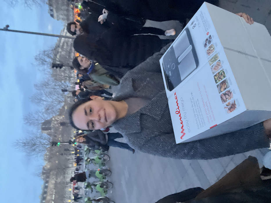 A woman holds a large white box in front of her, partially obscuring her face. The box is for a "Mouliner Easy Fry Mega" air fryer, displaying the product and images of cooked food. The woman is in an outdoor setting with other people and bicycles visible in the background. Bare trees and buildings suggest a city location, possibly in Europe. The lighting indicates it is either early morning or late afternoon.