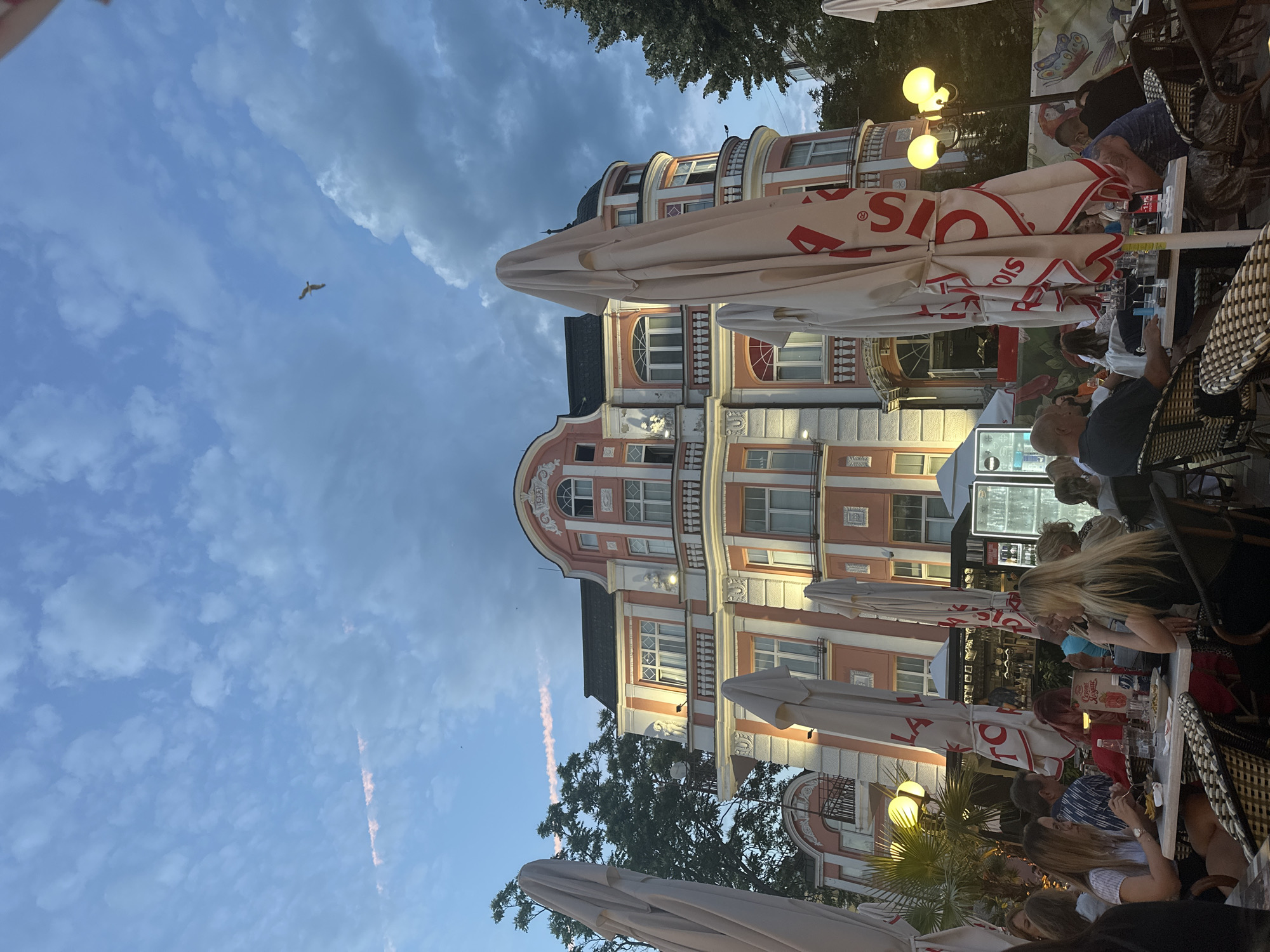 The image depicts an outdoor seating area of a restaurant or cafe in Varna, Bulgaria, situated in front of a grand, pink and white building with classical architectural elements. The sky above is a dusky blue, with scattered clouds and a contrail faintly visible. A lone bird is captured in flight against the sky. In the foreground, several large, white, closed umbrellas, some bearing red lettering that partially reads "LAZAROV," are prominent. Patrons are seated at tables, some of whom are eating and interacting. The overall ambiance suggests an evening setting, likely during or shortly after sunset, as evidenced by the lighting from street lamps and within the building.