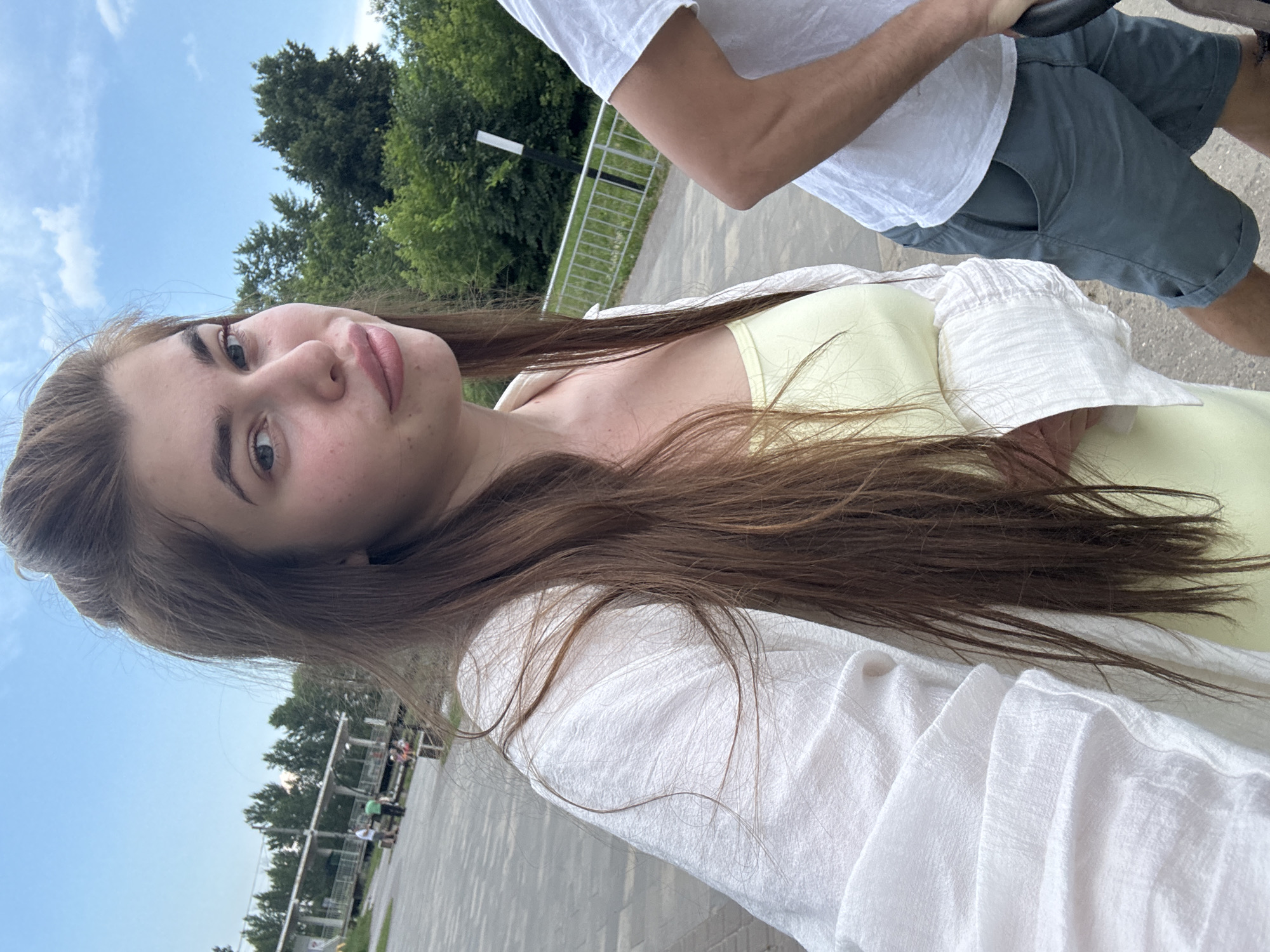 A young woman with long brown hair is taking a selfie outdoors. She is wearing a white open shirt over a light yellow top. Her hair is styled in a high ponytail. Her lips are slightly parted. Behind her, a man in a white t-shirt and grey shorts is partially visible to the right. The background shows a paved walkway, a metal fence, trees, and a cloudy blue sky. In the distance, there is a covered structure and several people. The location appears to be a park or recreational area in the city of Rechitsa, Russia.