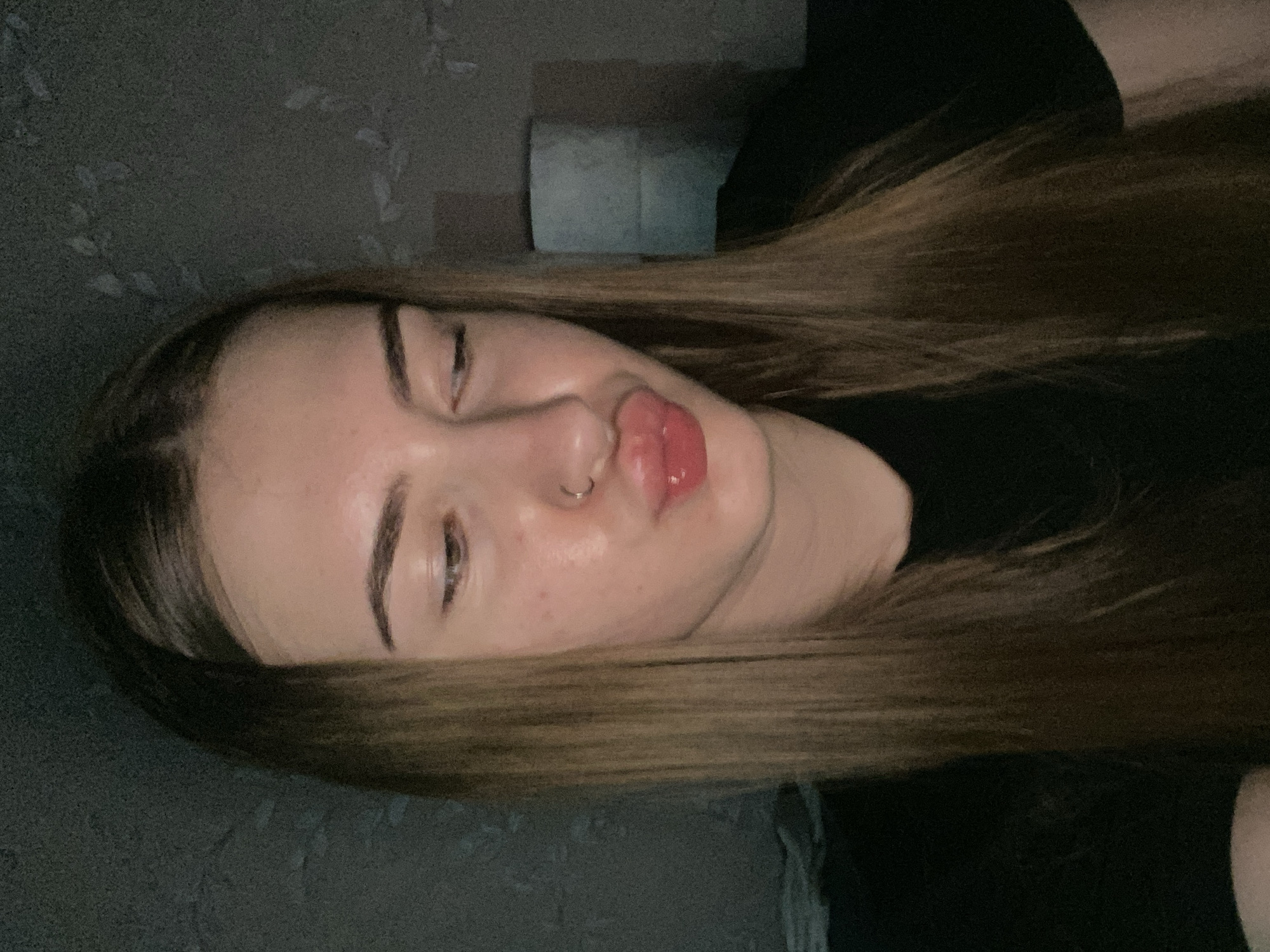 This is a close-up, head-and-shoulders, indoor portrait of a young woman with long, straight brown hair. She is wearing a black t-shirt and has a silver nose ring. Her lips are pursed and slightly glossy, and she is looking down and to her right. The background is dark and textured, with some faint, lighter patterns that could be wallpaper. A cylindrical object, possibly a roll of toilet paper or a lamp, is visible in the background to the right of the woman's head. The lighting is dim and appears to be artificial, creating shadows and a slightly grainy texture in the image. There is no visible indication of the location being in the city of Дьяково, Russia.