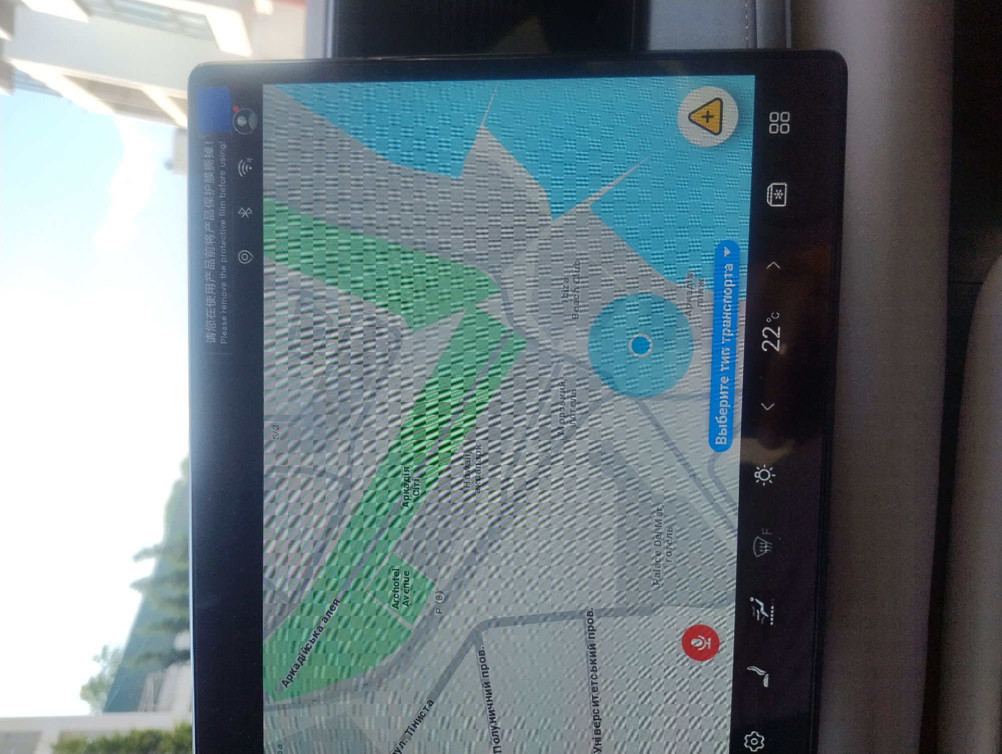 This media file is a vertical, close-up shot of a car's infotainment system screen, displaying a map navigation interface. The screen shows a portion of a city map, with green areas indicating parks or other open spaces and grey lines representing roads. Several street names and landmarks are visible, including "Аркадійська алея" (Arkadiyska Alley), "вул. Тіниста" (Tynysta St.), "Полуничний пров." (Polunychy Prow.), "Archotel Avenue", "Аркадія" (Arkadyia), "Hawait аквапарк" (Hawait Aquapark), and "Palace Del Mar готель" (Palace Del Mar Hotel).
On the right side of the screen, there are several icons related to navigation and vehicle settings, such as a temperature display showing "22°c", a traffic icon, a Wi-Fi symbol, and a settings gear. A prominent yellow triangular warning icon with a plus sign is also visible on the map.
Along the left edge of the screen, in white text, there is a message in both Chinese and English: "请您在使用产品前将产品保护膜撕掉! Please remove the protective film before using!". This suggests the device is new and has a protective film that needs to be removed.
The background of the image, seen through the car's windshield on the left, is bright and appears to be daytime with clear skies, suggesting it's likely sunny. The overall setting is the interior of a vehicle, with the focus solely on the illuminated screen. There are no people visible. The image has a slightly grainy or pixelated appearance, possibly due to the camera quality or the screen's display technology.