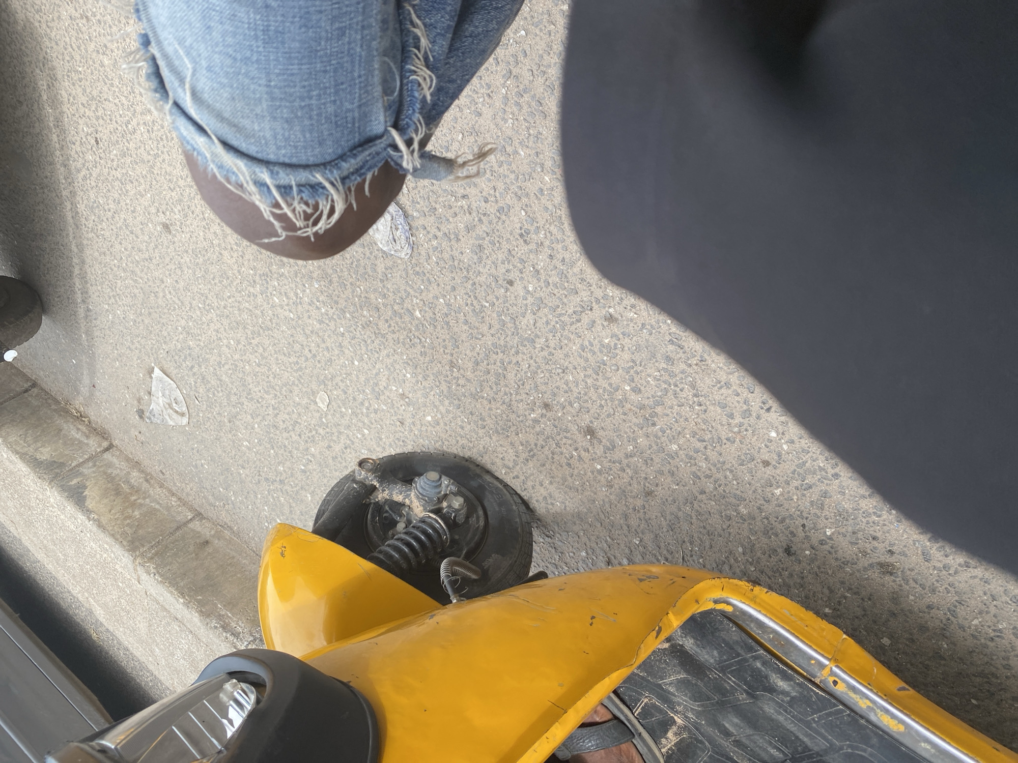 This is a high-angle, close-up shot of a person's legs and a yellow electric scooter on an asphalt surface, likely a street or sidewalk. The person is wearing distressed denim jeans with frayed hems and dark sandals. The scooter, predominantly yellow, shows signs of wear and tear with some scratches and scuffs visible on its body. The front wheel and part of the suspension system are visible. The ground is a textured asphalt with small pebbles. To the left, a concrete curb separates the asphalt from a lighter gray surface. Shadows suggest it is daytime, with sunlight casting distinct shapes on the ground. The overall impression is that of an urban outdoor setting, possibly in Jalingo, Nigeria, where someone is standing next to their scooter. No visible text is present in the image.