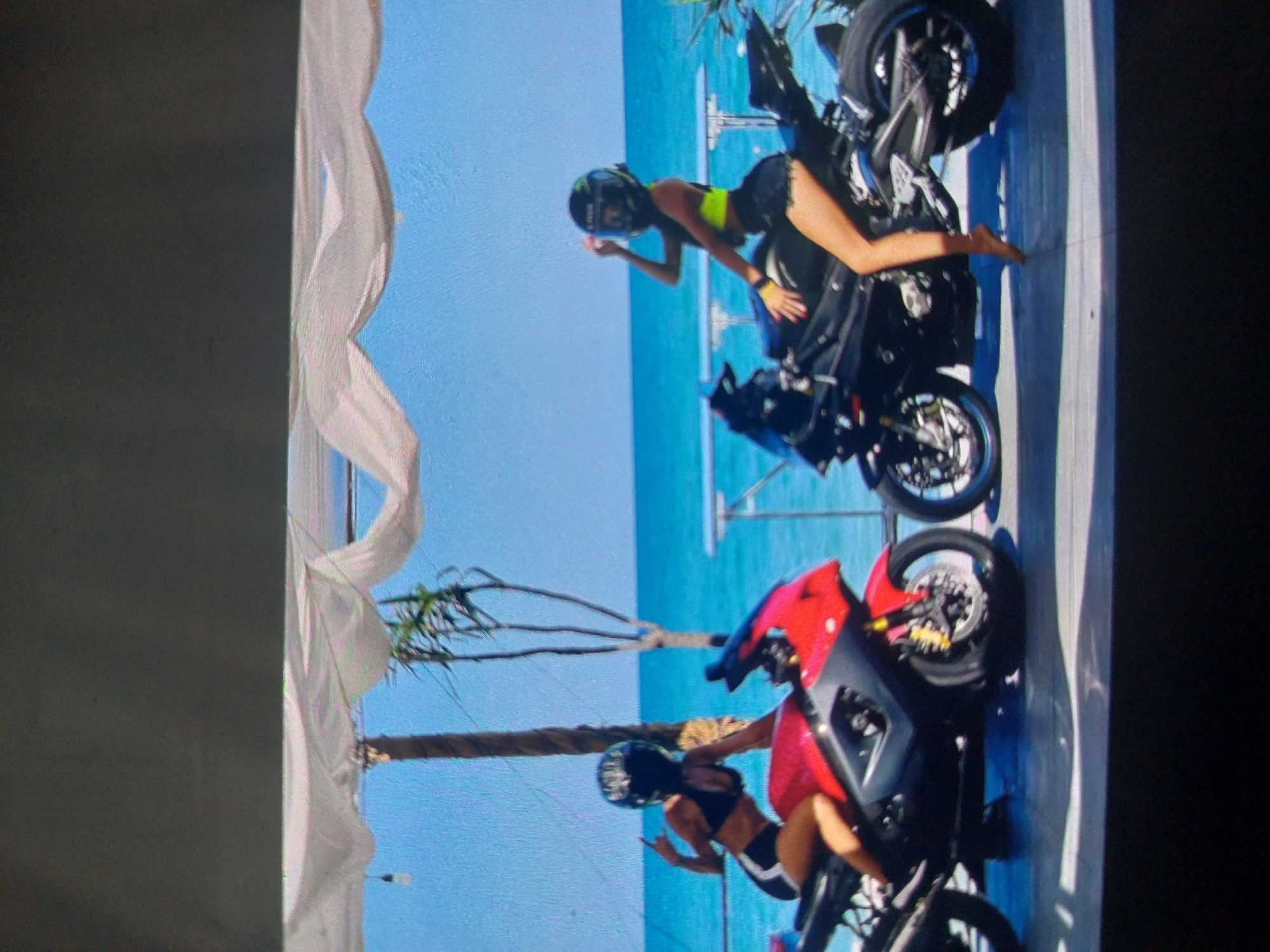 The image depicts a coastal scene, presumably in the city of Tsarevo, Bulgaria, given the prompt. Two women wearing helmets are positioned on motorcycles against the backdrop of a beach and turquoise sea.
The first woman, wearing a neon-yellow top and dark shorts, is partially reclined on a black motorcycle. Her bare foot rests on the frame of the bike. The motorcycle faces to the left.
Below and slightly to the left, the second woman, wearing a black top and dark shorts, is seated on a red motorcycle. Her posture and position suggest she is also posing. The motorcycle faces to the left.
The scene is partially framed by what appears to be white drapery and palm trees. The overall environment is bright and sunny, typical of a coastal resort.