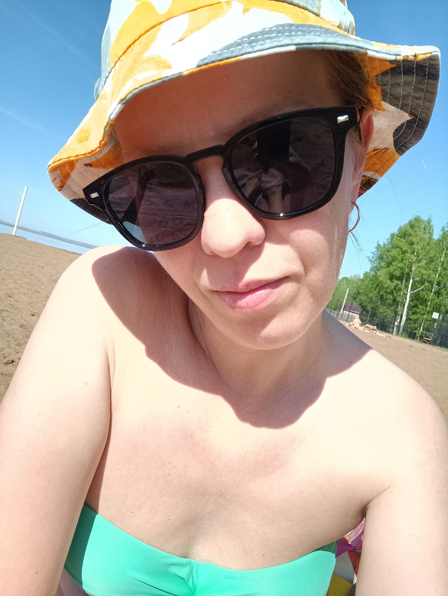 A woman is taking a selfie on a beach. She is wearing a yellow and grey patterned bucket hat and dark sunglasses. She is also wearing a green bathing suit top. The beach appears to be sandy, with some trees and a building in the background. The sky is blue. The location is Chaikovsky, Russia.