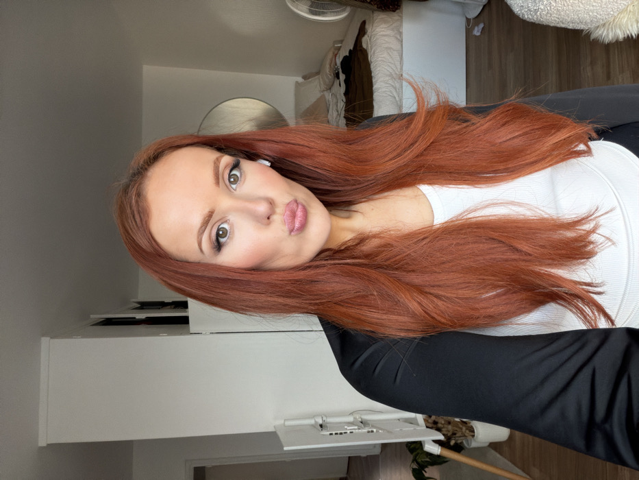 The scene is an interior space located in Paris, France. A female subject, presenting with long, wavy reddish-brown hair and light skin, is depicted from the upper torso, directly facing the viewer. She is dressed in a white ribbed tank top worn under a black long-sleeved top. Her lips are pursed, and a white wireless earbud is present in her left ear. The environment consists of white walls. To the subject's left, a white vertical stand featuring horizontal elements, possibly a monitor arm or shelving unit, stands next to a potted plant containing dried botanicals. A partial view of a wooden furniture leg and an open doorway leading to another room are also visible. To the subject's right, a bed with white linens and a brown throw is present, with some garments resting upon it. A round mirror with a thin metallic frame hangs on the wall above the bed. A white stand fan is situated on the far right. A white wall-mounted structure is partially visible near the top right corner. The room is brightly illuminated.