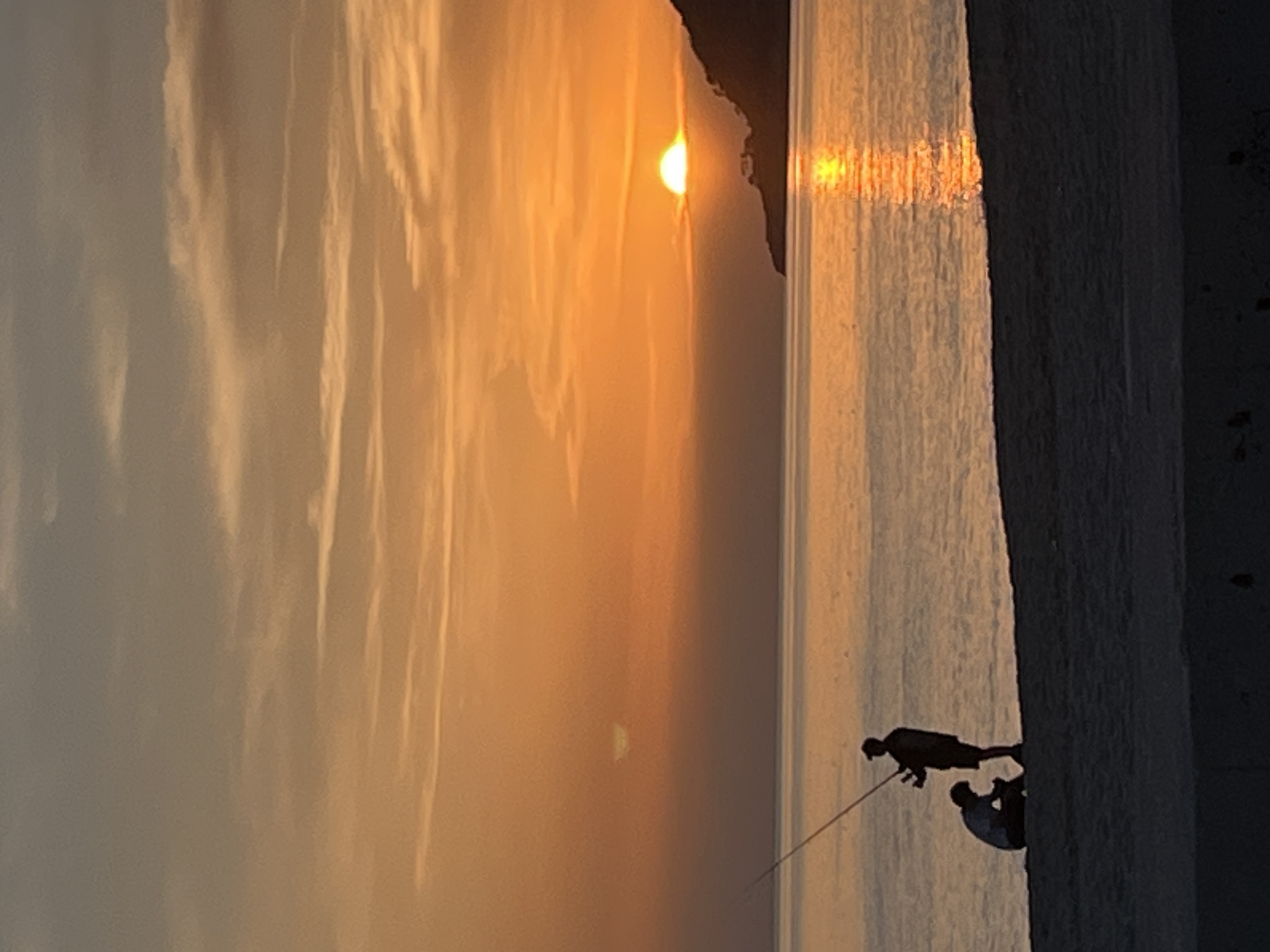 The image captures a sunset scene in Alanya, Turkey. The sun is positioned near a rocky headland. Reflections of sunlight stretch across the water's surface. Two individuals are silhouetted at the edge of the water, one appearing to hold a fishing rod. The sky displays a gradient of orange and light. A structure, potentially a wall or cliff face, is visible in the foreground.