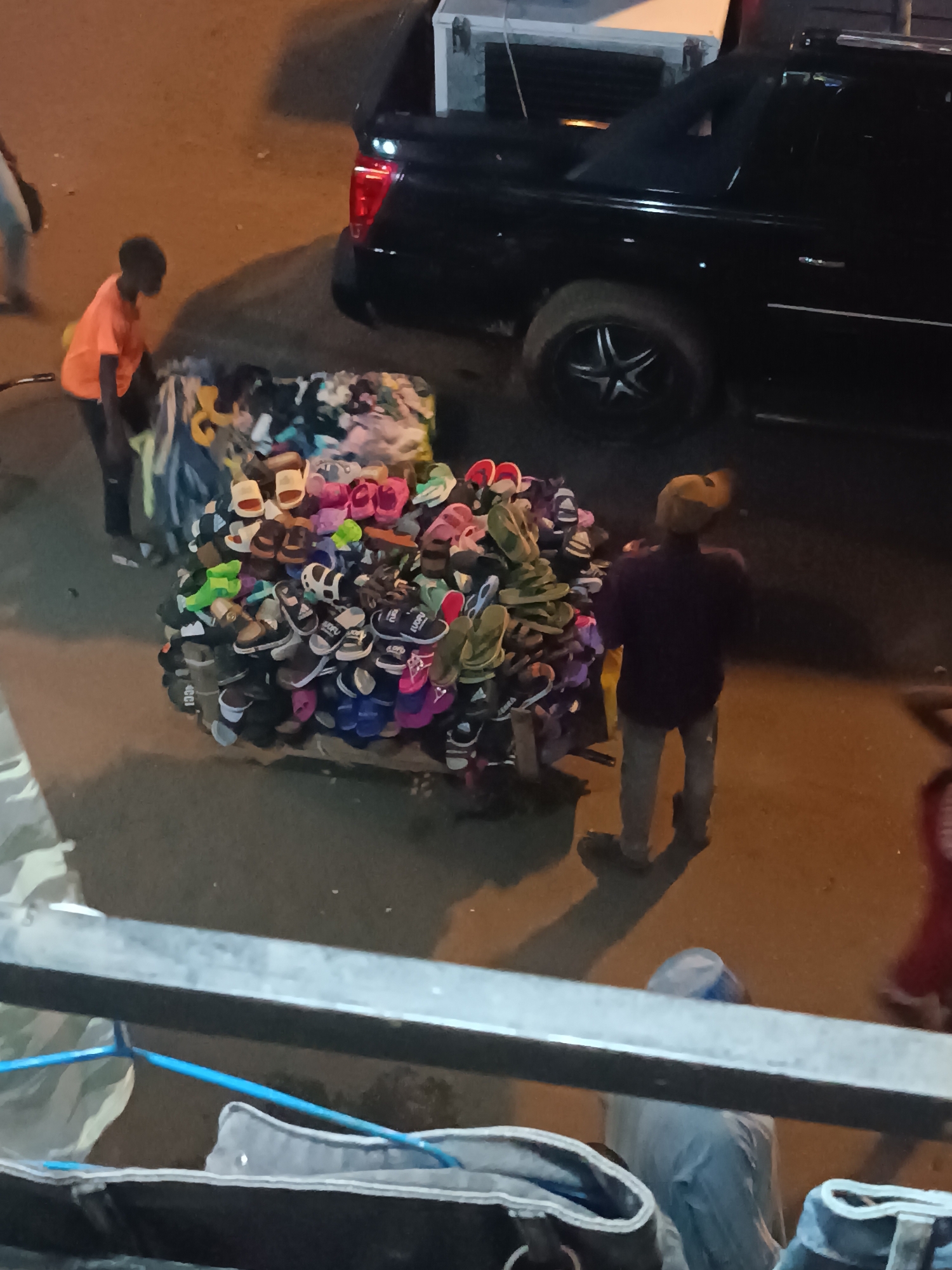 In Bukuru, Nigeria, an outdoor scene shows two individuals near a large assortment of shoes piled on a wooden structure, potentially for sale. One individual, wearing an orange shirt, is on the left, appearing to arrange or move the shoes. Another person, wearing a dark shirt and lighter pants with a head covering, stands to the right, facing the shoes. A black vehicle with its trunk open is parked in the background. The lighting suggests it is either evening or a dimly lit area. The scene is viewed from an elevated position.