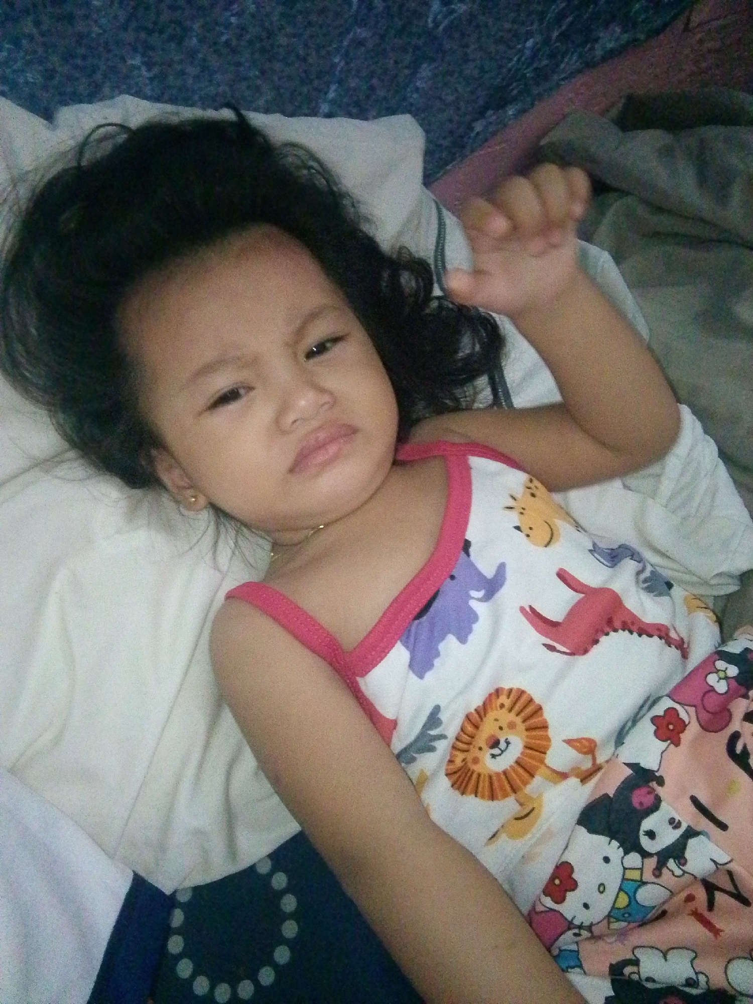 A young East Asian or Southeast Asian child is depicted lying down on a white sheet, presenting a slight frown or pout. The child has dark, disheveled hair fanned around the head. A reddish mark is visible on the forehead. The child is wearing a white sleeveless top with pink trim, featuring cartoon animal illustrations including a purple elephant, a red dinosaur, a yellow giraffe, and an orange lion. Below the top, patterned pajama bottoms with Hello Kitty and My Melody characters are visible. A small gold-colored stud earring is present in the left ear, and a thin gold-colored necklace is visible around the neck. The child's left hand is partially visible, slightly clenched. The background consists of various textiles, including a dark blue/purple textured fabric on the upper left, a muted grey/brown fabric on the upper right, and a dark blue/black fabric with a pattern of light grey circles below the child's lower body. The scene is indoors.