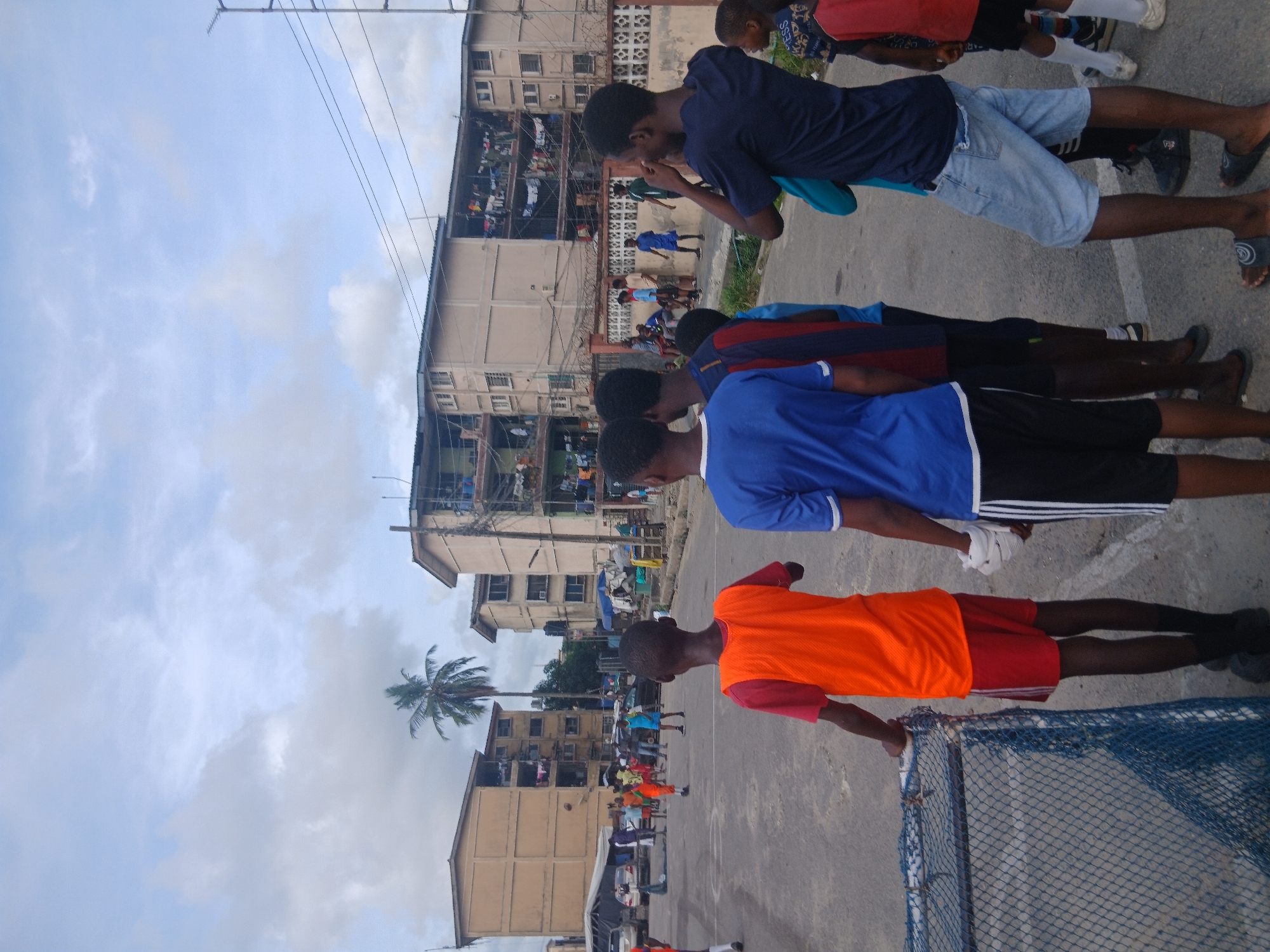 A group of young men, mostly of African descent, stand on an outdoor basketball court in Lagos, Nigeria. The first man wears an orange shirt and red shorts, standing by a basketball net. Behind him, two men wear blue shirts and dark shorts. Farther back, another man wears a blue shirt and denim shorts. Several buildings are nearby, where clothing hangs.