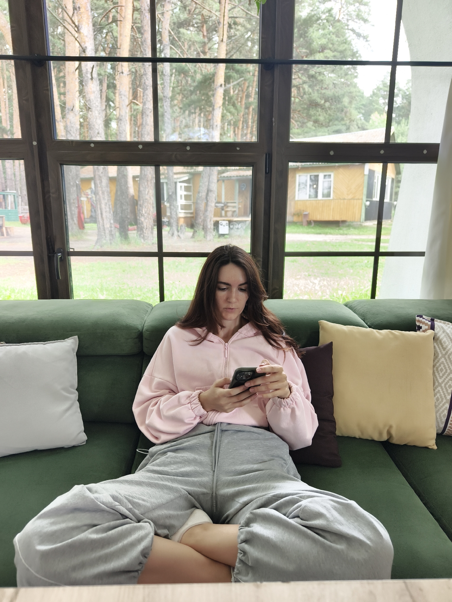 A young woman with long, dark brown hair is seated on a green sofa, facing the camera. She is wearing a light pink zip-up hoodie and loose-fitting gray sweatpants. Her legs are spread apart, and she is looking down at her black smartphone, which she holds in both hands. The sofa is adorned with several throw pillows: a large white pillow on the left, a dark brown pillow, and two patterned pillows on the right, one yellow and one with a geometric design.
Behind the woman is a large window with dark brown wooden frames, divided into panes. Through the window, a wooded area with tall pine trees is visible. Several small, yellow, wooden buildings with white trim and windows are scattered among the trees. The ground outside appears to be covered in green grass. The setting suggests a rural or suburban environment, possibly a dacha or a resort area, consistent with locations like Onochino, Russia, where such structures are common. The lighting is natural and diffused, indicating it is daytime.