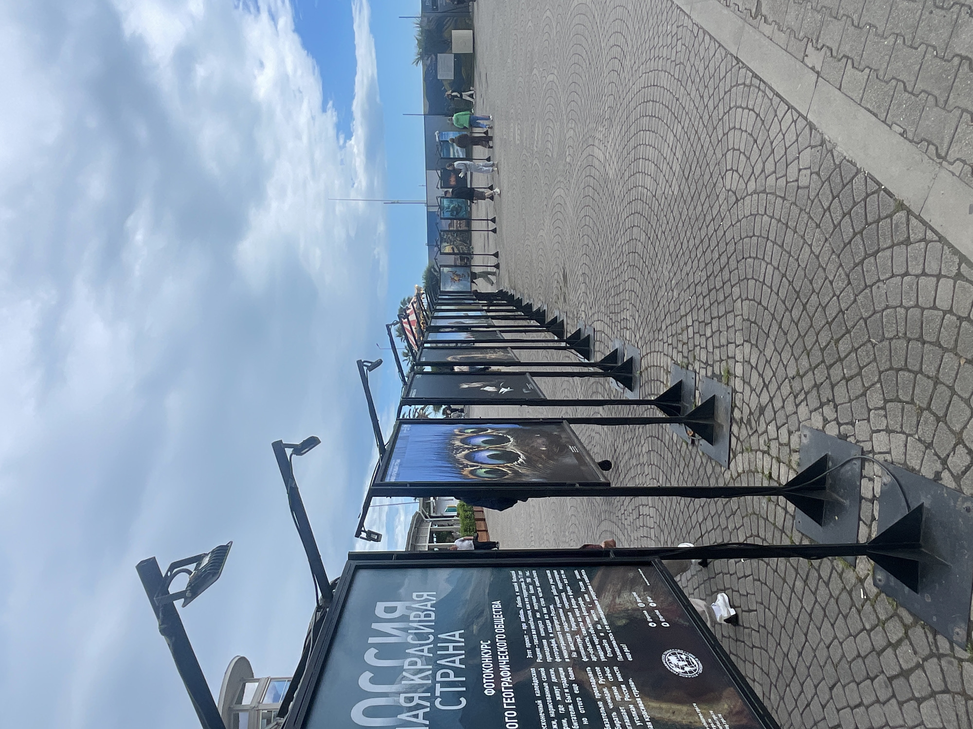 The image shows an outdoor exhibition in Sochi, Russia, showcasing photographs from the Russian Geographical Society's photo contest. The display is arranged along a paved walkway, with each photograph mounted on a black stand. The background features a partly cloudy sky and some buildings and people further down the walkway. The text on the display reads "Russia is a beautiful country".