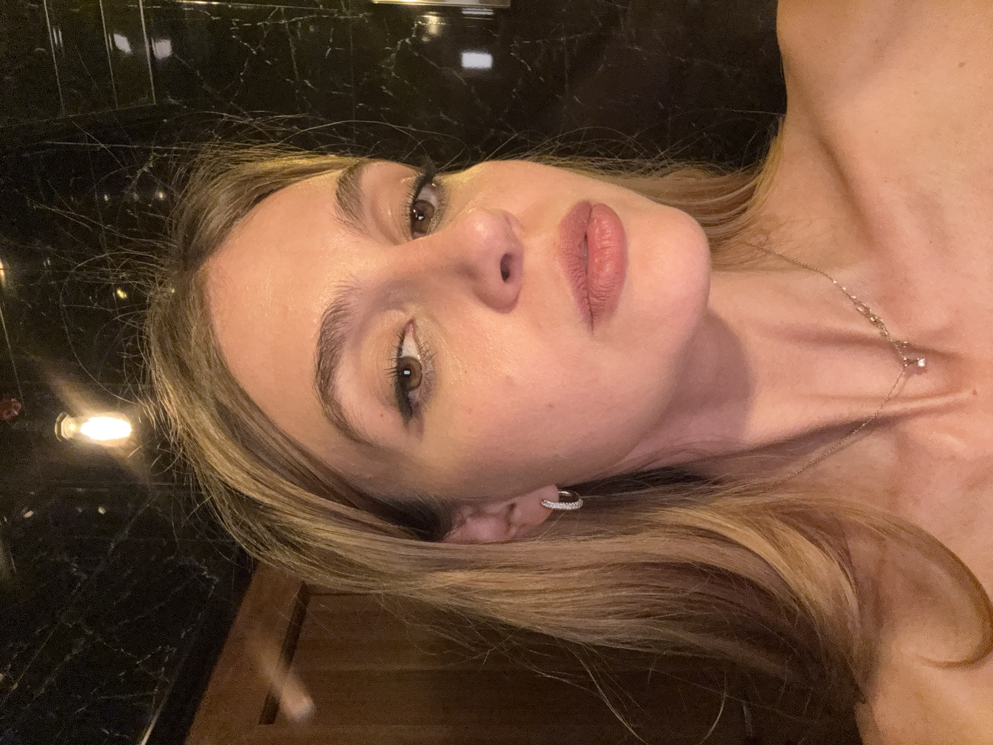 A woman with fair skin, light brown hair, and brown eyes is the primary subject of this image. She is wearing an earring in her left ear and a delicate necklace. The background features what appears to be a dark, polished surface, possibly marble, with light reflections. The location is not identifiable as Yerevan, Armenia, based on the visual information provided.