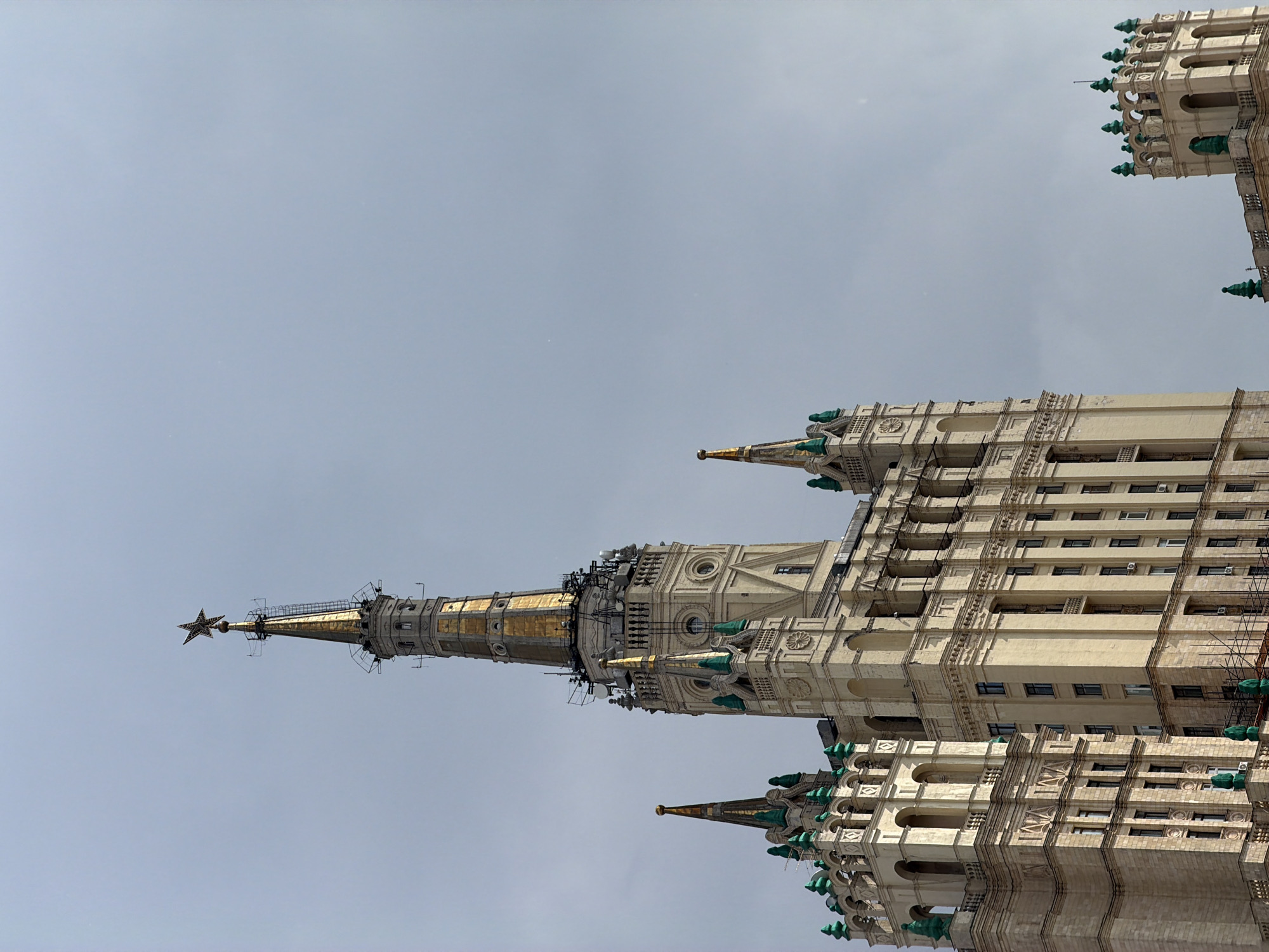 The image shows a low-angle view of the Kotelnicheskaya Embankment Building, one of the Seven Sisters skyscrapers in Moscow, Russia. The building has a prominent spire topped with a star. The sky is overcast. The building is ornate with multiple tiers and decorative elements, including small spires with green roofs. The building is multi-storied, with many windows. The image appears to be taken from street level, looking upward.