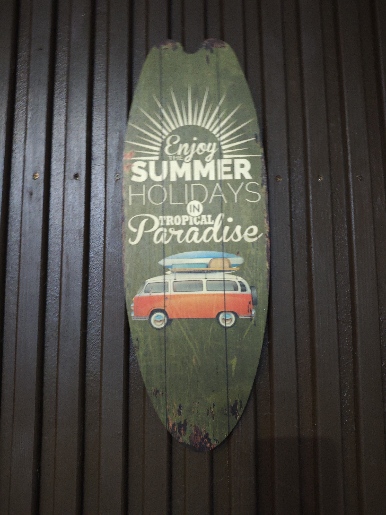 The image shows a decorative surfboard-shaped sign hanging on a dark, vertically-paneled wall. The sign is predominantly green with the words "Enjoy the Summer Holidays in Tropical Paradise" written in white and cream-colored fonts. Above the text, there's a stylized sunburst graphic. Below the text, an image of a red and white vintage Volkswagen van with surfboards strapped to the roof is visible. The sign has a distressed appearance with spots of what appears to be simulated wear and tear. The background wall has vertical grooves and a dark brown or black color. The location, Chaikovsky, Russia, cannot be determined from the image alone.