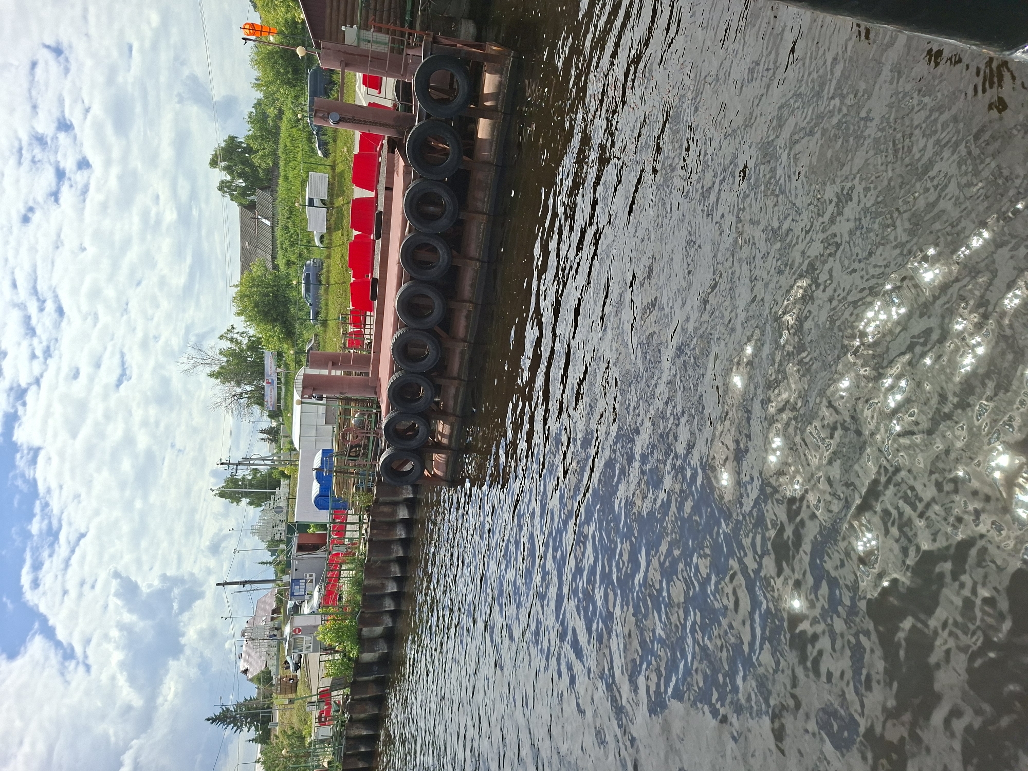 This is a medium-angle, eye-level shot of a ferry landing at the city of Verkhny Uslon, Russia. The ferry landing is made of metal and is lined with tires, which serve as bumpers. Red seats are visible on the ferry, and there are several structures behind the landing, including buildings and trees. The water in the foreground is rippled and reflects the sunlight. The sky is cloudy, with patches of blue visible. The image is taken on a sunny day.