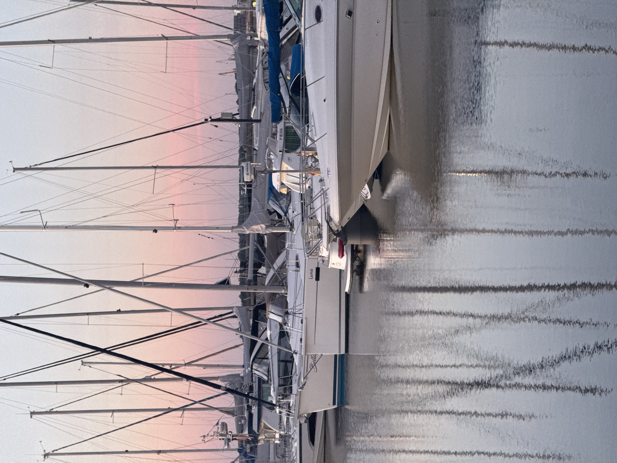 This is a photograph of sailboats docked in a harbor. The sky in the background is a pale pink and blue, suggesting either sunrise or sunset. The boats are white with tall masts and rigging, and they are reflected in the calm water. The scene is serene and peaceful. The location is not specified in the image.