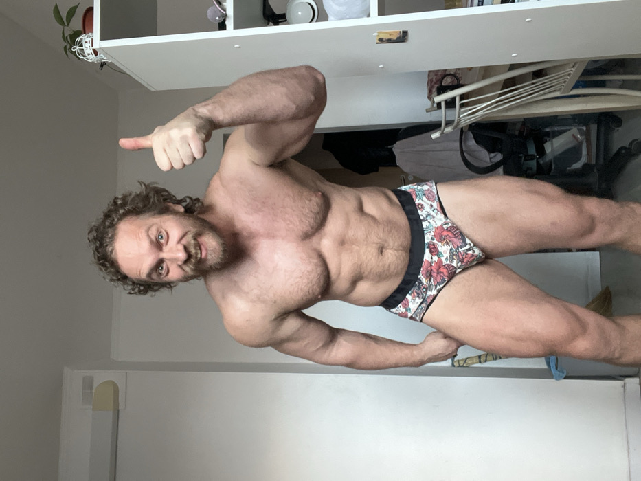 An adult male with curly brown hair and a beard stands bare-chested, wearing patterned briefs, in an indoor room in Saint Petersburg, Russia. The man faces forward, exhibiting a muscular physique, and is captured in a broad smile with wide eyes. His right hand is raised, forming a thumbs-up gesture. The background on the left consists of a plain, light-colored wall with a horizontal fixture mounted high. On the right, a white shelving unit is visible, displaying a small potted plant on its top shelf. A dark office chair draped with fabric and part of a light-colored chair are also present in the background.